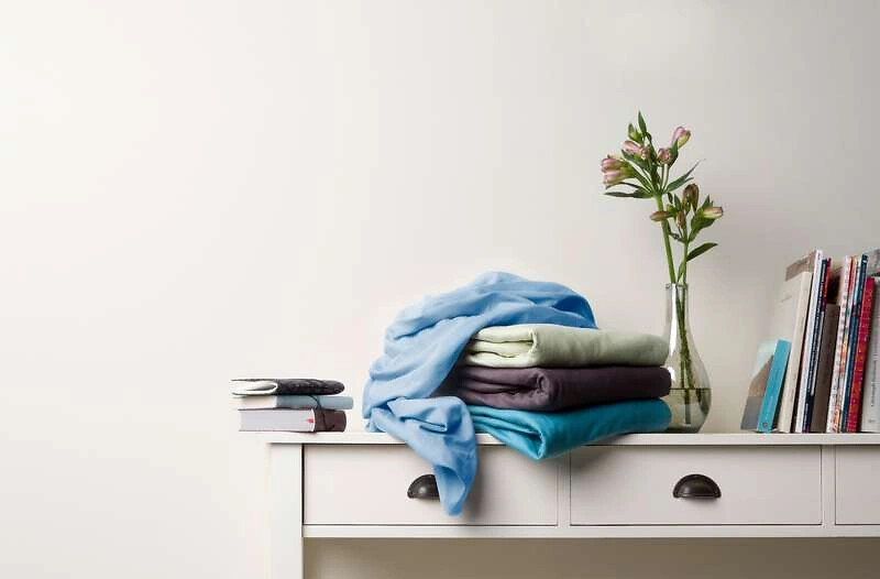 Gefaltete Spannbettlaken in verschiedenen Tönen auf weißer Kommode mit Vase und Büchern