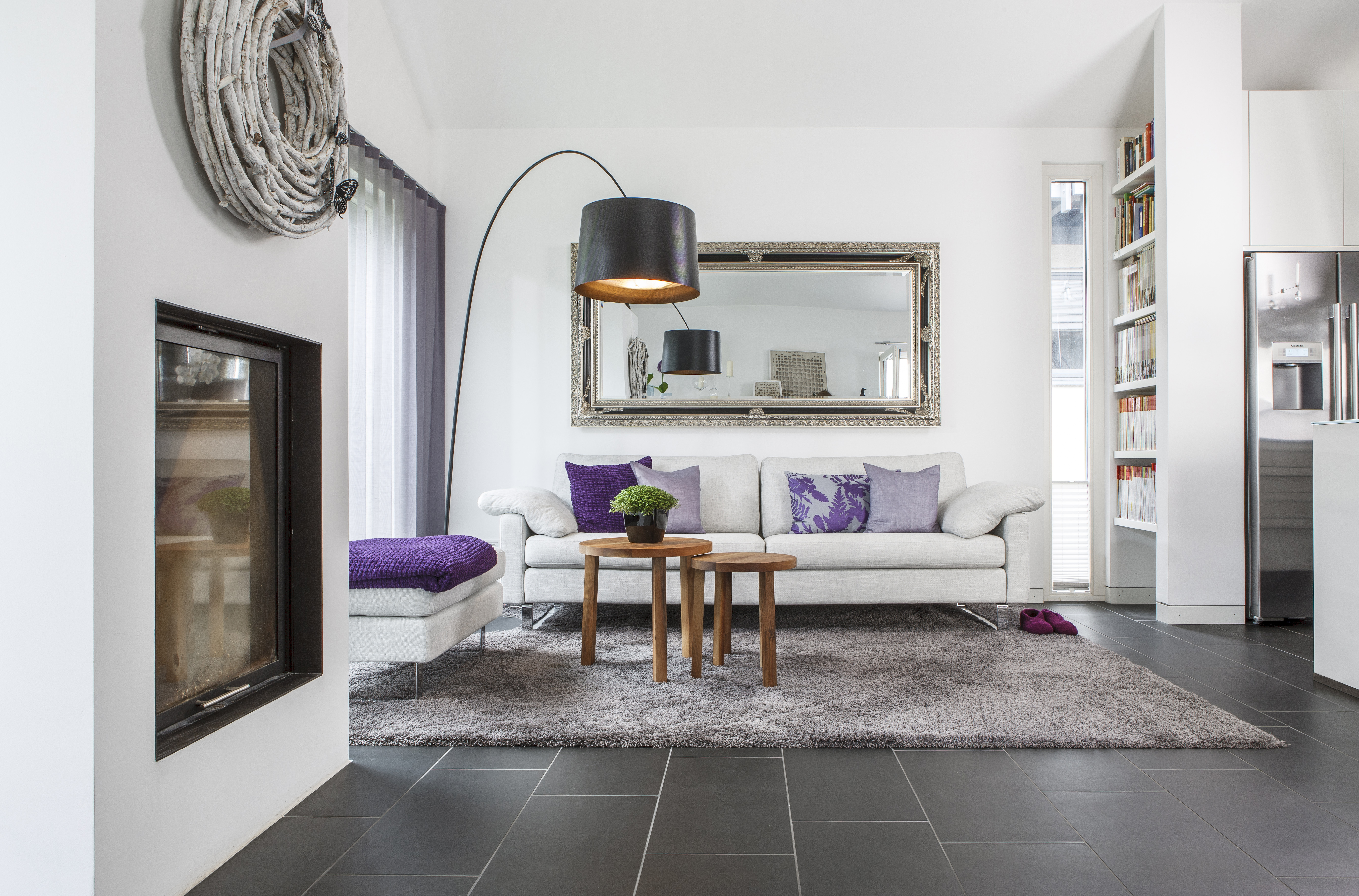 Wohnzimmer mit weißem Sofa, lila Akzenten und Spiegel über dem Sofa. Teppich auf dunklem Fliesenboden.