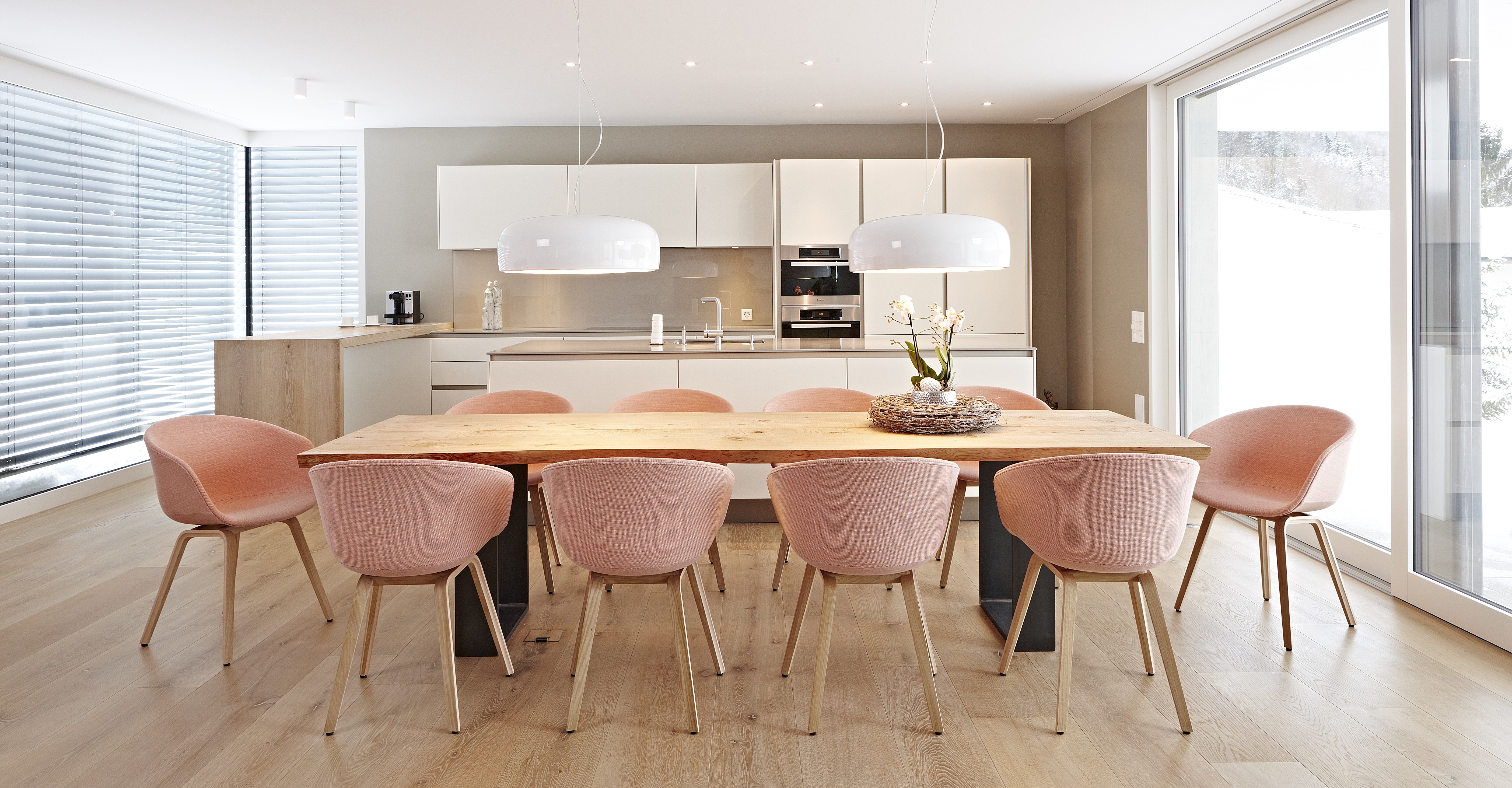 Großer Holztisch mit acht roséfarbenen Schalenstühlen, dahinter moderne Küche in hellen, matten Tönen.