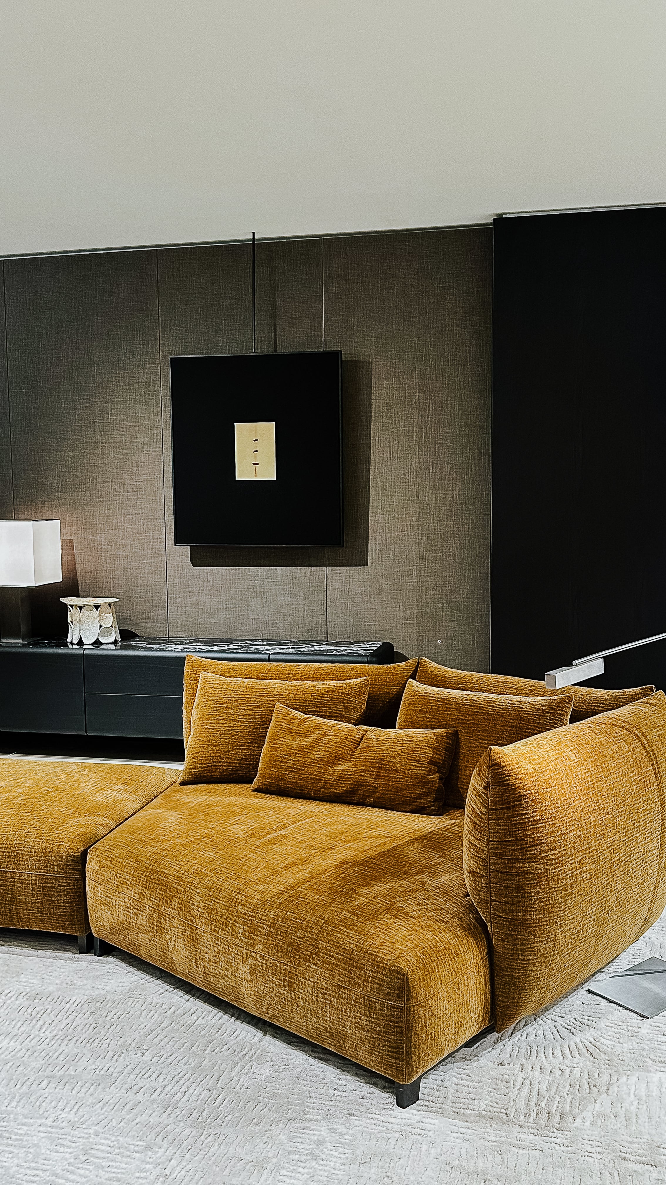 Ausstellungsansicht von der Il Salone del Mobile 2025 mit skulpturalem Sofa in Ockergelb, grauem Teppich und dunklem Sideboard. Im Hintergrund wandhängendes Kunstobjekt und stilisierte Leuchte.