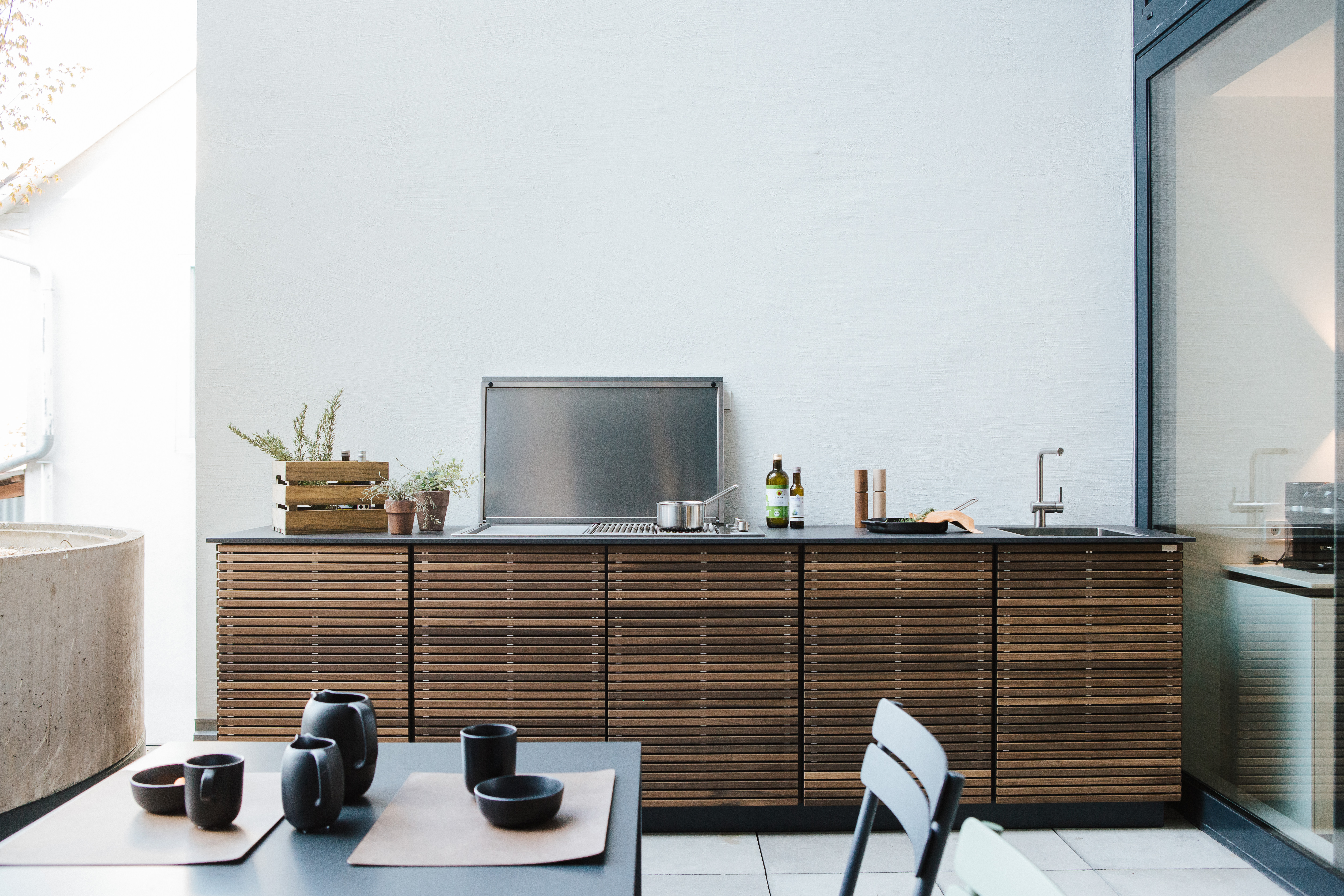 Modulare Outdoor-Küche von Cubic Outdoor Living mit Holzlamellenfront, integriertem Spülbecken und Gasgrill in einem lichtdurchfluteten Innenhof. Im Vordergrund ein Tisch mit schwarzem Geschirr.