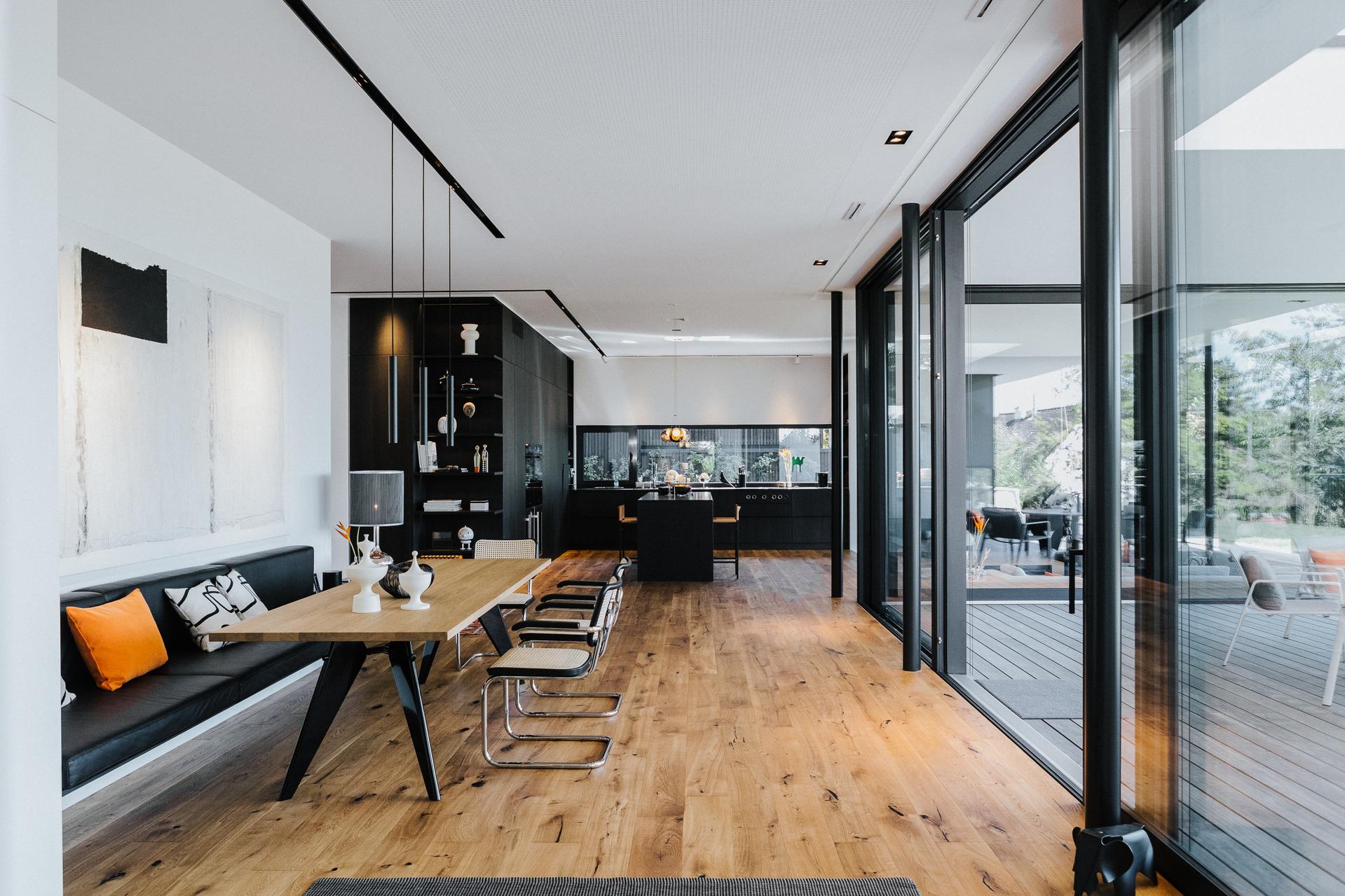 Offener Wohn- und Essbereich mit schwarzer Einbauküche, Holzboden, langer Massivholztisch, Vitra EM Table, mit Freischwinger-Stühlen, Thonet S64V und integrierter Sitzbank. Große Fensterfront mit Blick auf die Terrasse.