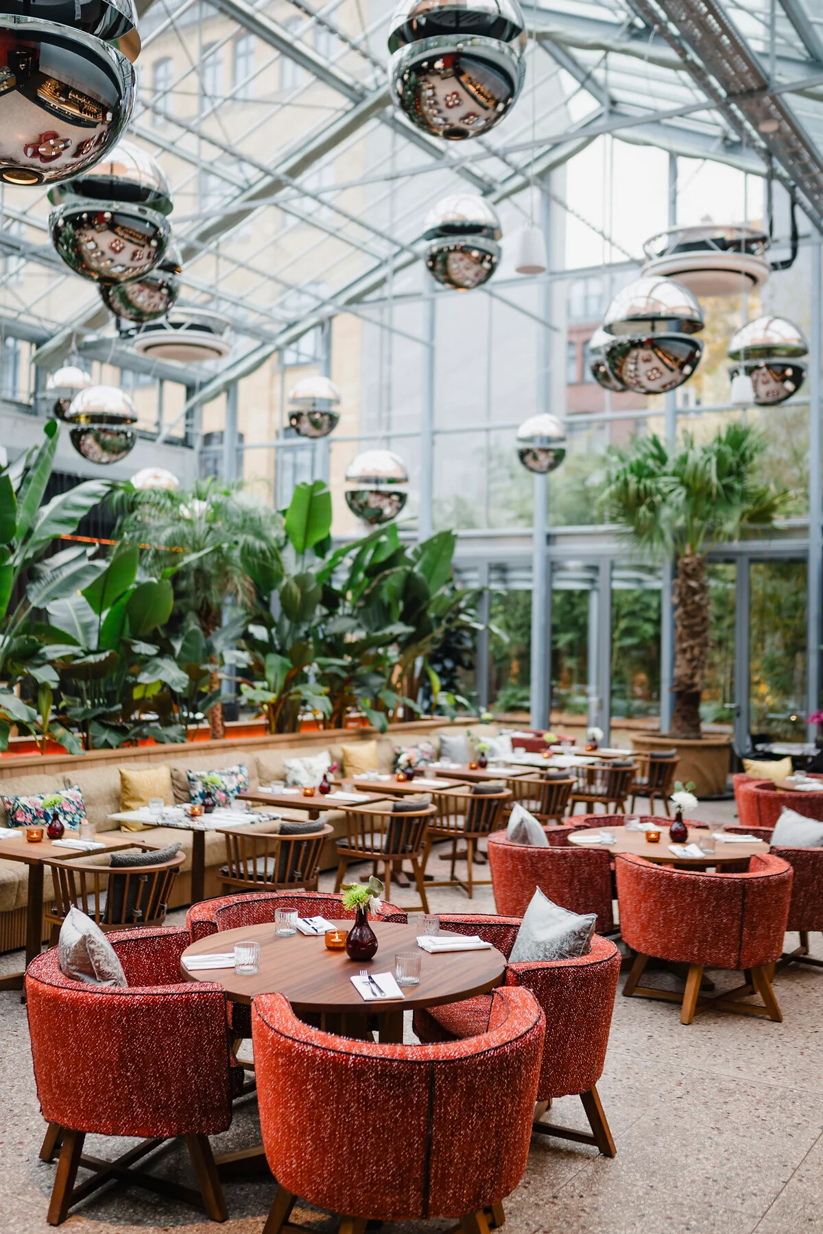 Runde Tische mit roten Sesseln in einem verglasten Gastraum mit Begrünung und Hängelampen.