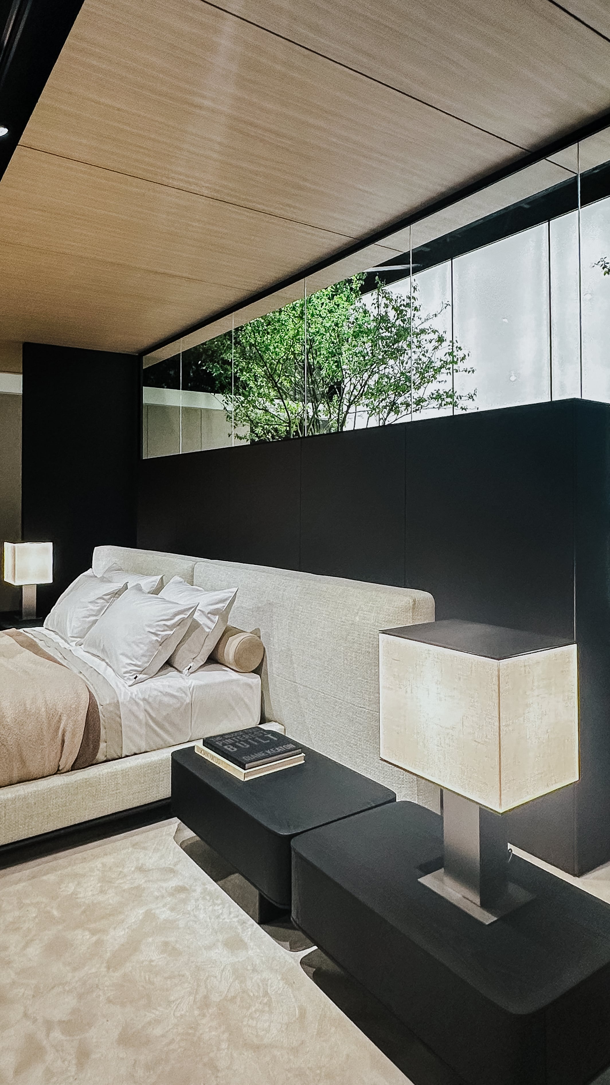 Messeinstallation auf der Il Salone del Mobile 2025 mit Polsterbett, schwarzen Beistelltischen und markanten Leuchten. Raum mit Holzdecke, Teppichboden und Fensterband mit Blick ins Grüne.