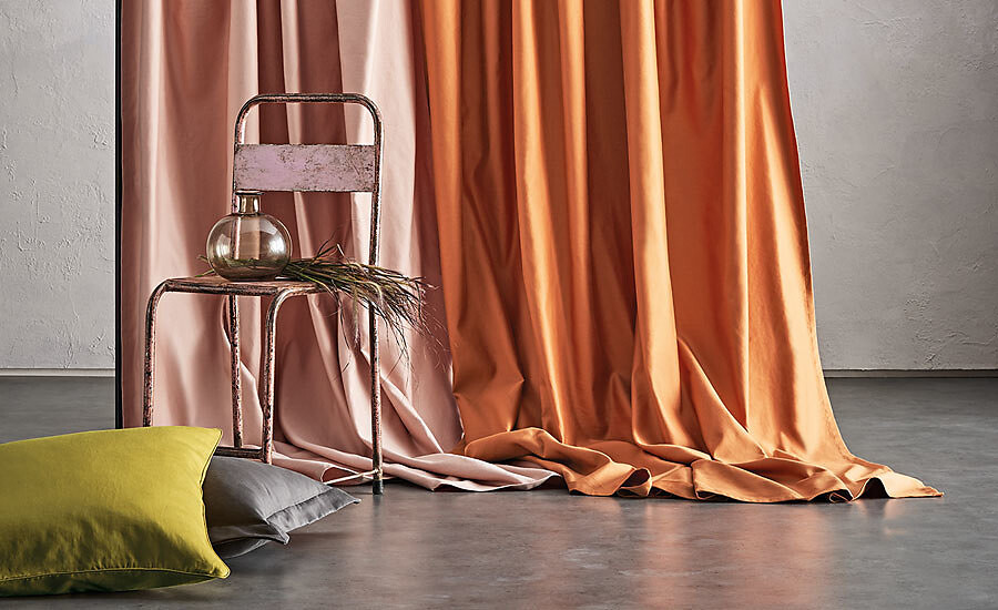 Schwere Vorhänge in Orange und Rosé auf grauem Boden neben Metallstuhl und Kissen