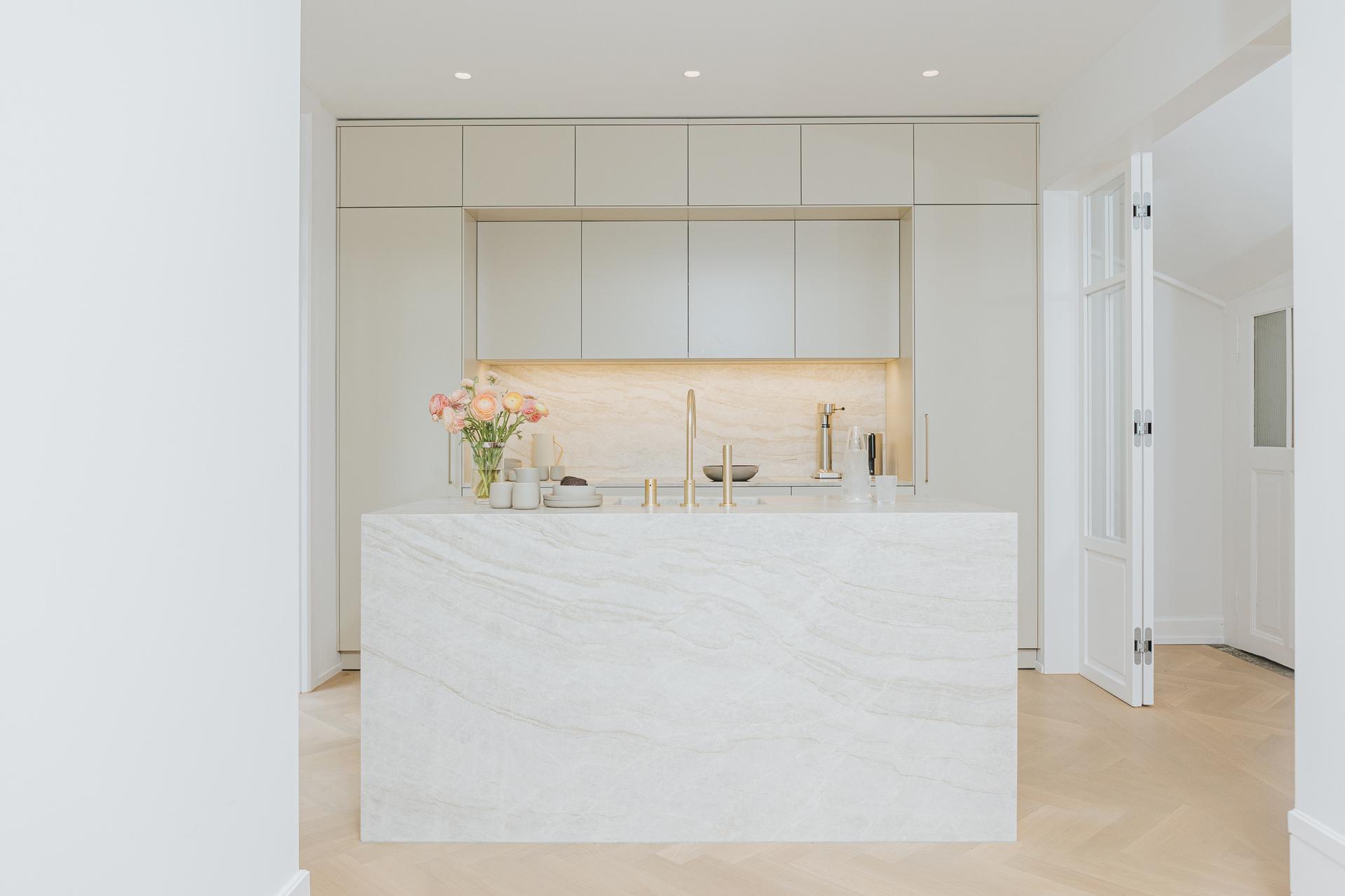 Minimalistische Küche in hellem Ton mit Kücheninsel aus Naturstein, goldener Armatur und Vase mit Blumen.