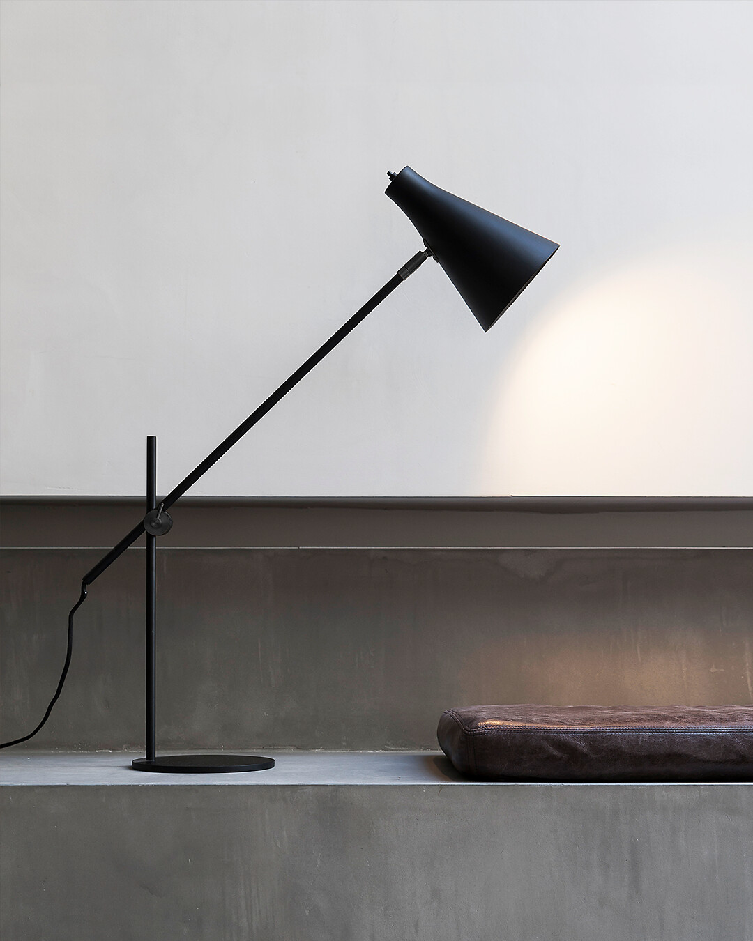 Schwarze Tischleuchte mit geneigtem Schirm auf grauer Ablage vor heller Wand