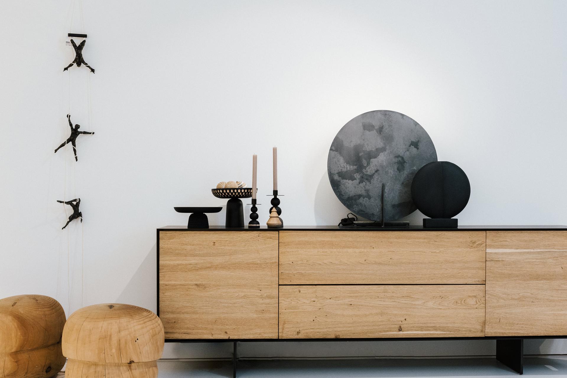 Sideboard aus Holz mit dekorativen Objekten vor weißer Wand, daneben Hocker und Wandfiguren.