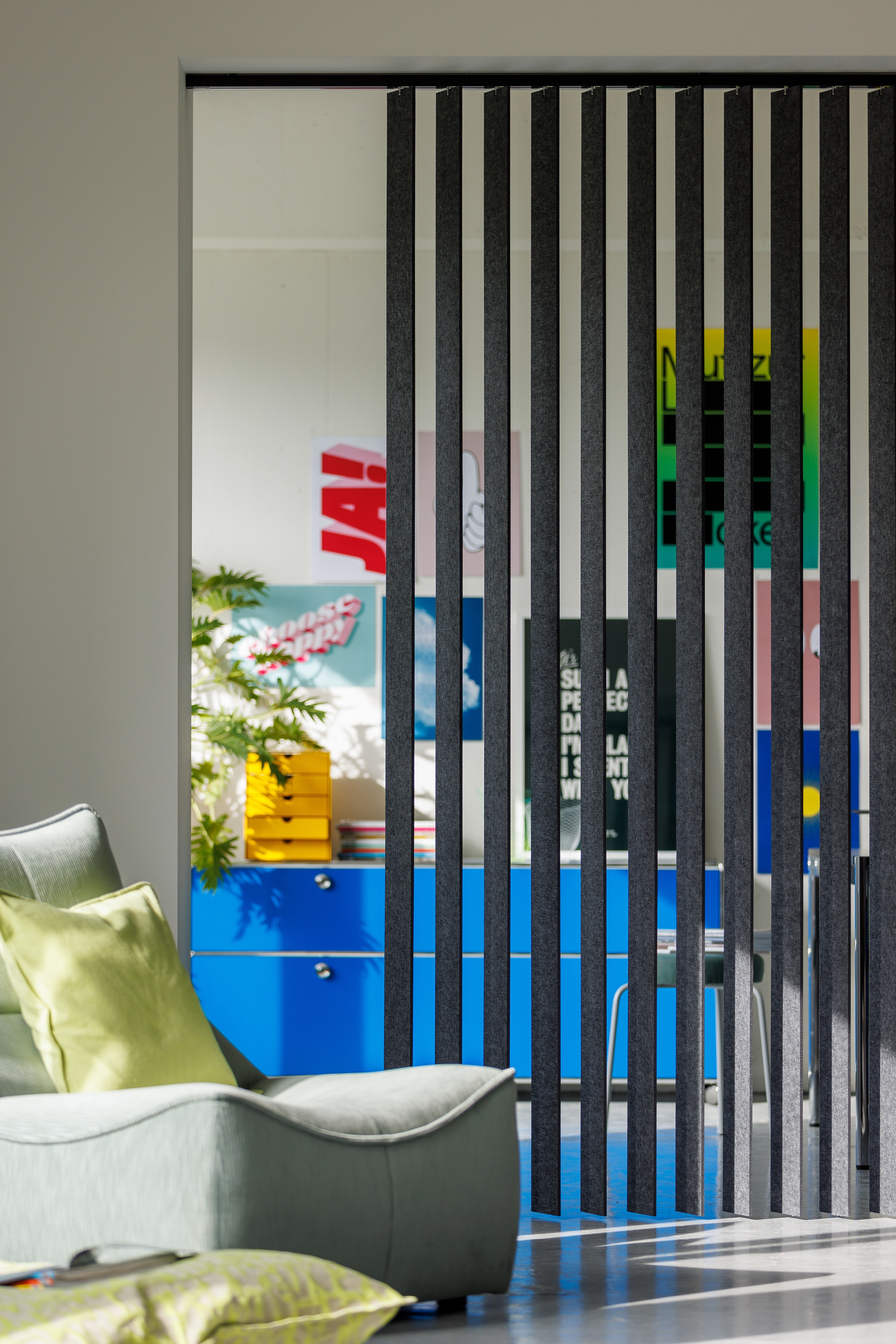 Raumteiler aus vertikalen Filzlamellen vor bunt eingerichteter Wohnlandschaft mit Sessel