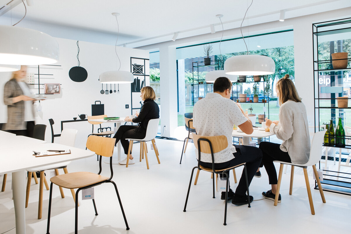 Menschen im Café Super Normal an weißen Tischen in heller Umgebung mit großen Fenstern und Regalwand.