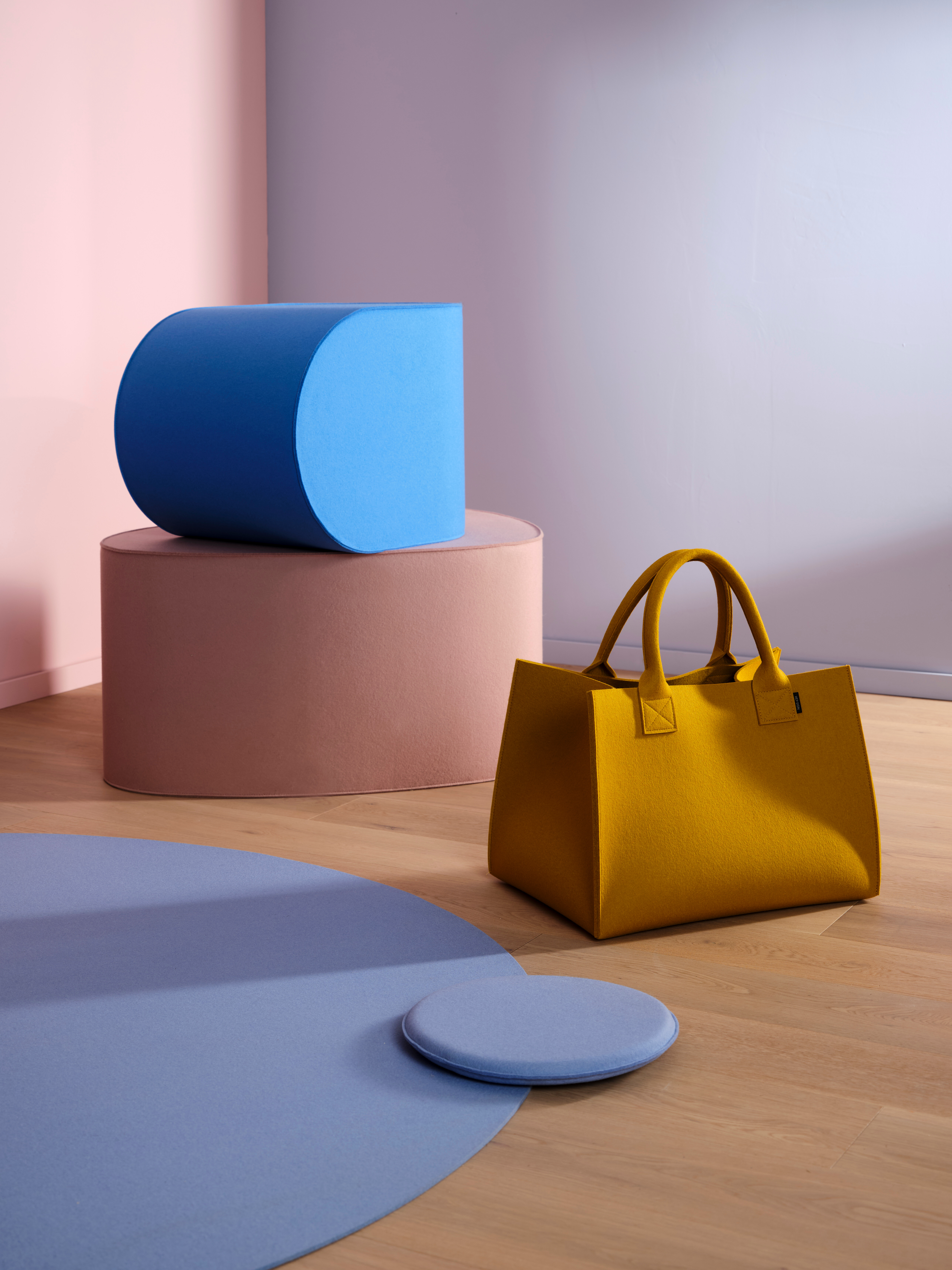 Senffarbene Filztasche vor pastellfarbenen Formen und blauem Sitzkissen