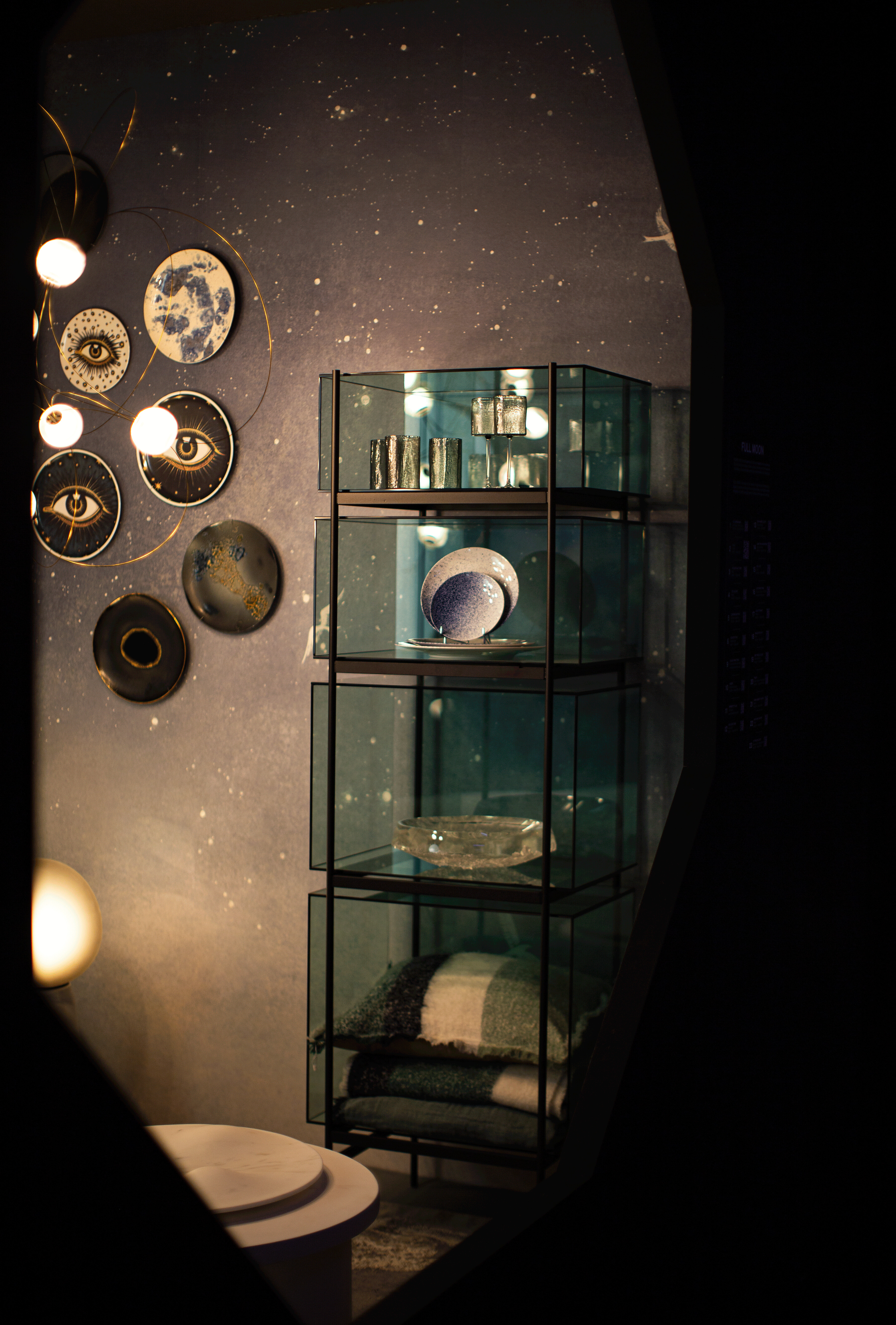 Vitrinenschrank aus Glas mit Tellern, Gläsern und Stoffen vor dunkler Wand mit Dekor