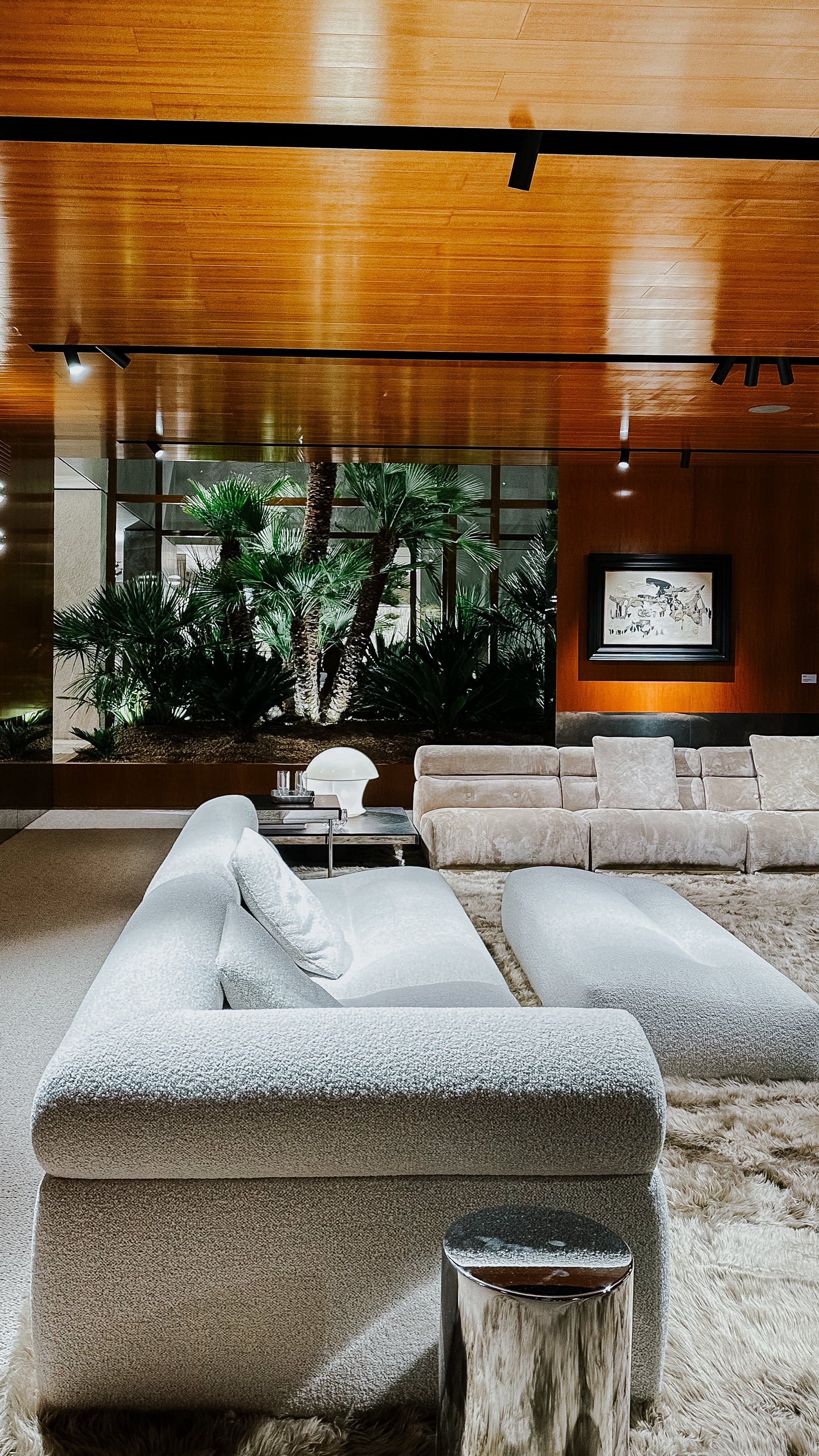 Sofalandschaft auf der Il Salone del Mobile 2025 in weißem Bouclé, kombiniert mit hochflorigem Teppich und verchromtem Beistelltisch. Raumkulisse mit Holzdecke, tropischer Begrünung und kunstvoller Wandgestaltung.