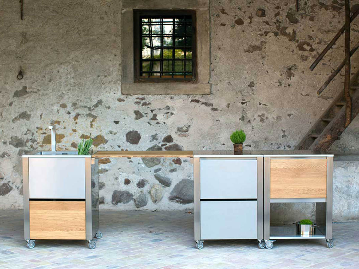 Modulare Outdoorküche von Jokodomus mit Edelstahl- und Holzelementen vor rustikaler Steinwand, verbunden durch eine Holzplatte.