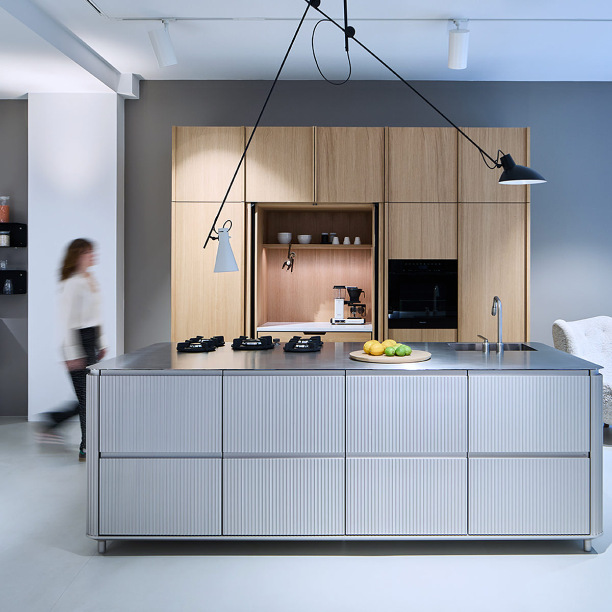 Showroom: Vipp V3 Küche, Symbiose aus Funktionalität & Design. Edelstahl-Kücheninsel nahtlos verbunden mit maßgefertigtem Eichenfurnier-Hochschrank, dessen Pocket Doors Ausstattung elegant verbergen.