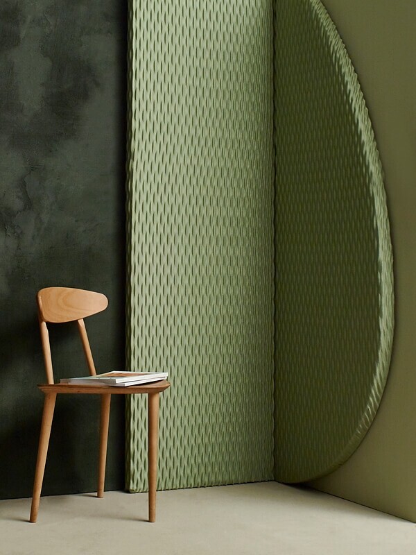 Grüne Akustikpaneele mit feiner Textur an der Wand, davor Holzstuhl mit Buch