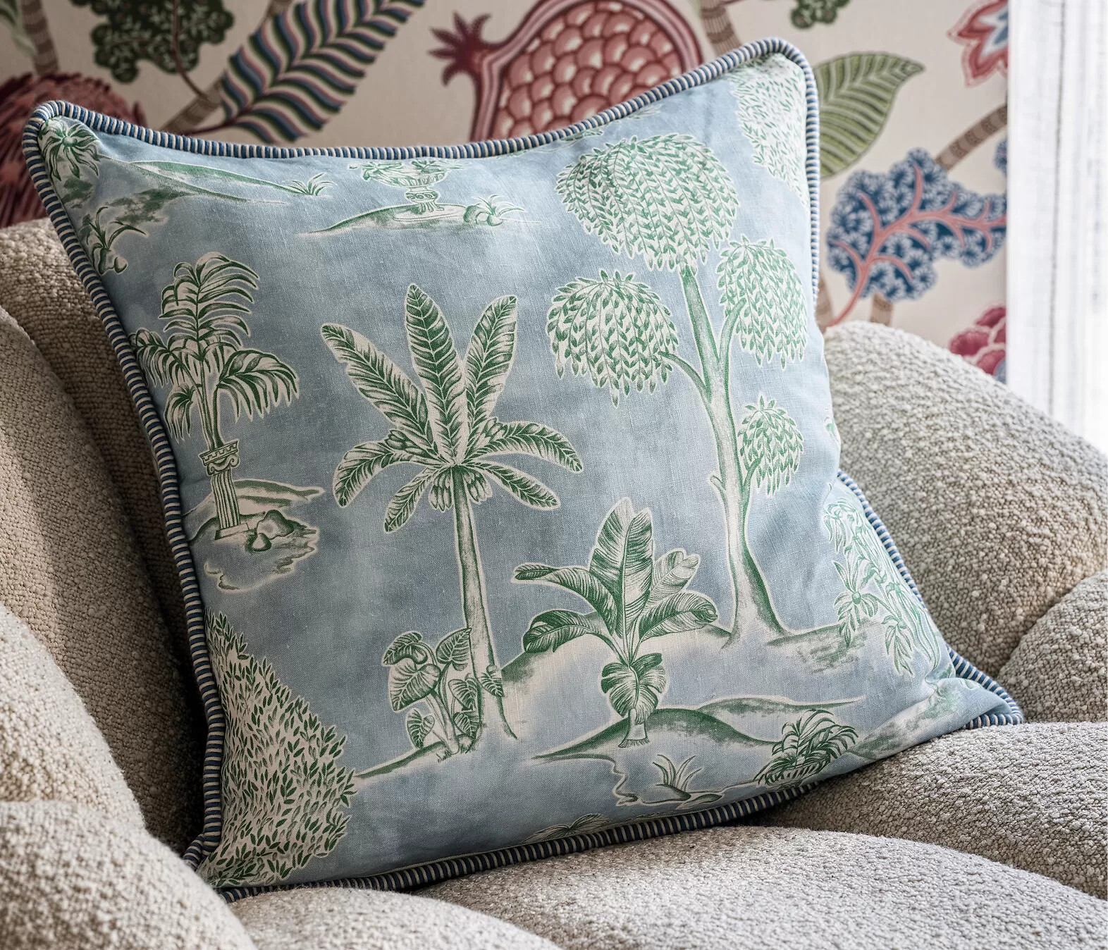Dekokissen mit Palmenmotiv auf beigefarbenem Bouclé-Sofa vor gemusterter Tapete