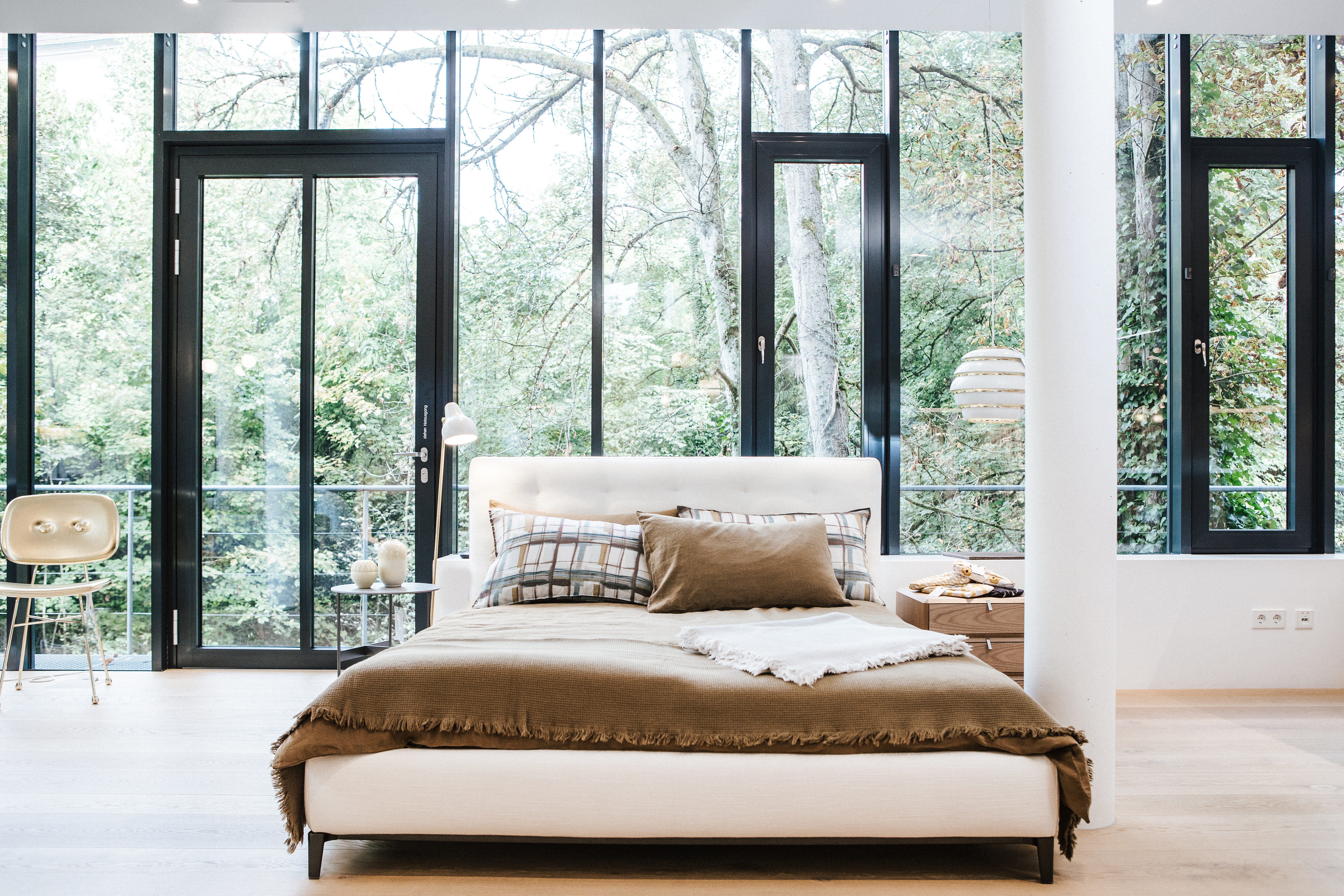 Schlafzimmer mit hellem Polsterbett vor bodentiefer Fensterfront mit Blick ins Grüne