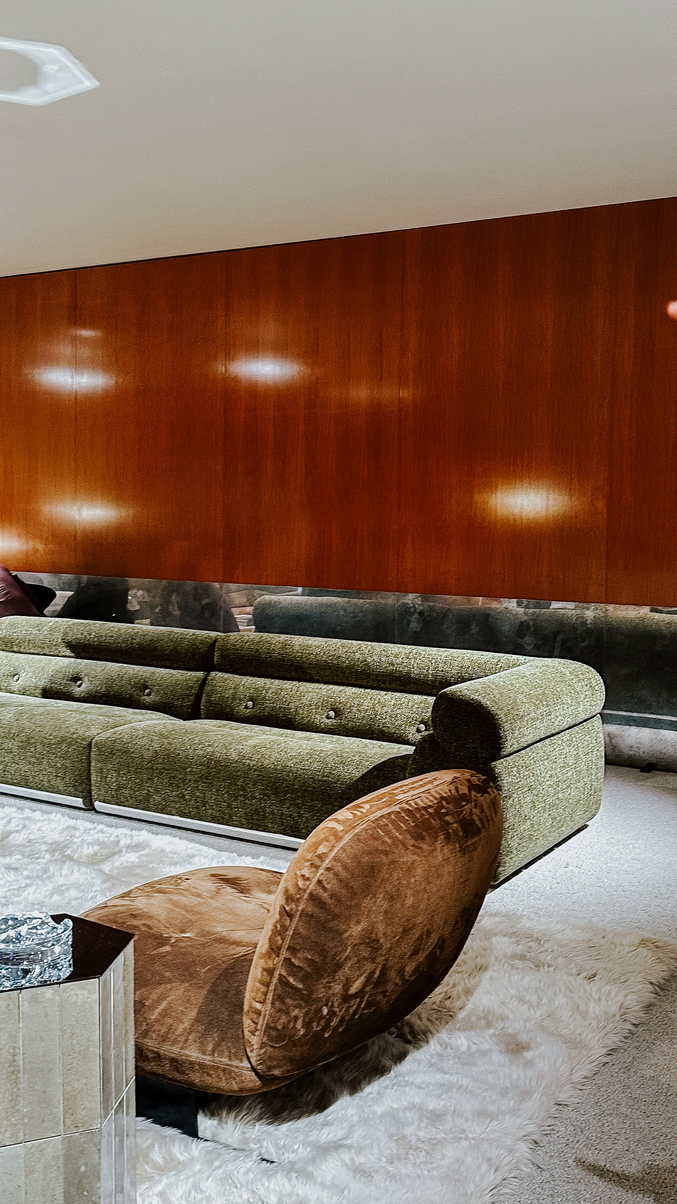 Sofainszenierung auf der Il Salone del Mobile 2025 mit grüner Polstergruppe, rundem Loungesessel in Braun und hochglänzendem Beistelltisch. Die Wand ist mit dunklem Holz verkleidet, der Boden mit hellem Teppich ausgelegt.