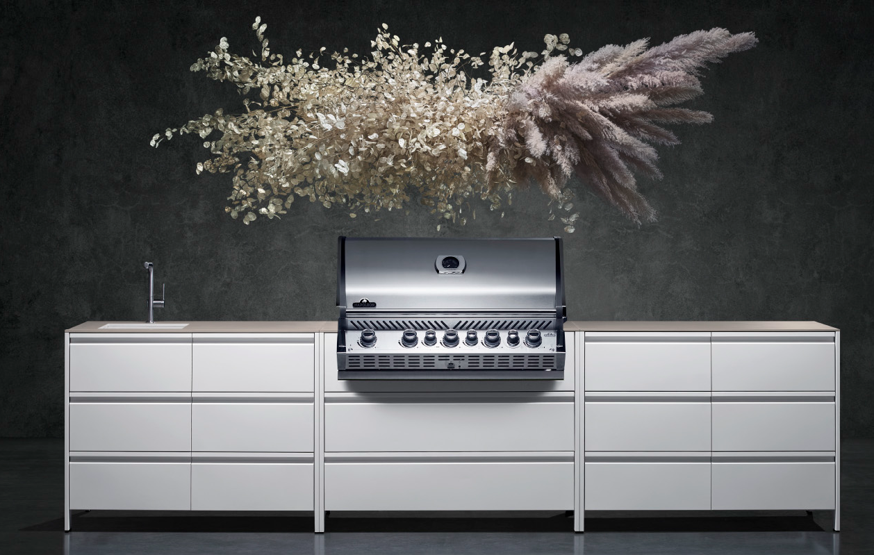 Modulare Belmento Outdoor-Küche mit Gasgrill und Spüle, symmetrisch angeordnet vor dunkler Wand und dekorativem Trockenblumen-Arrangement.