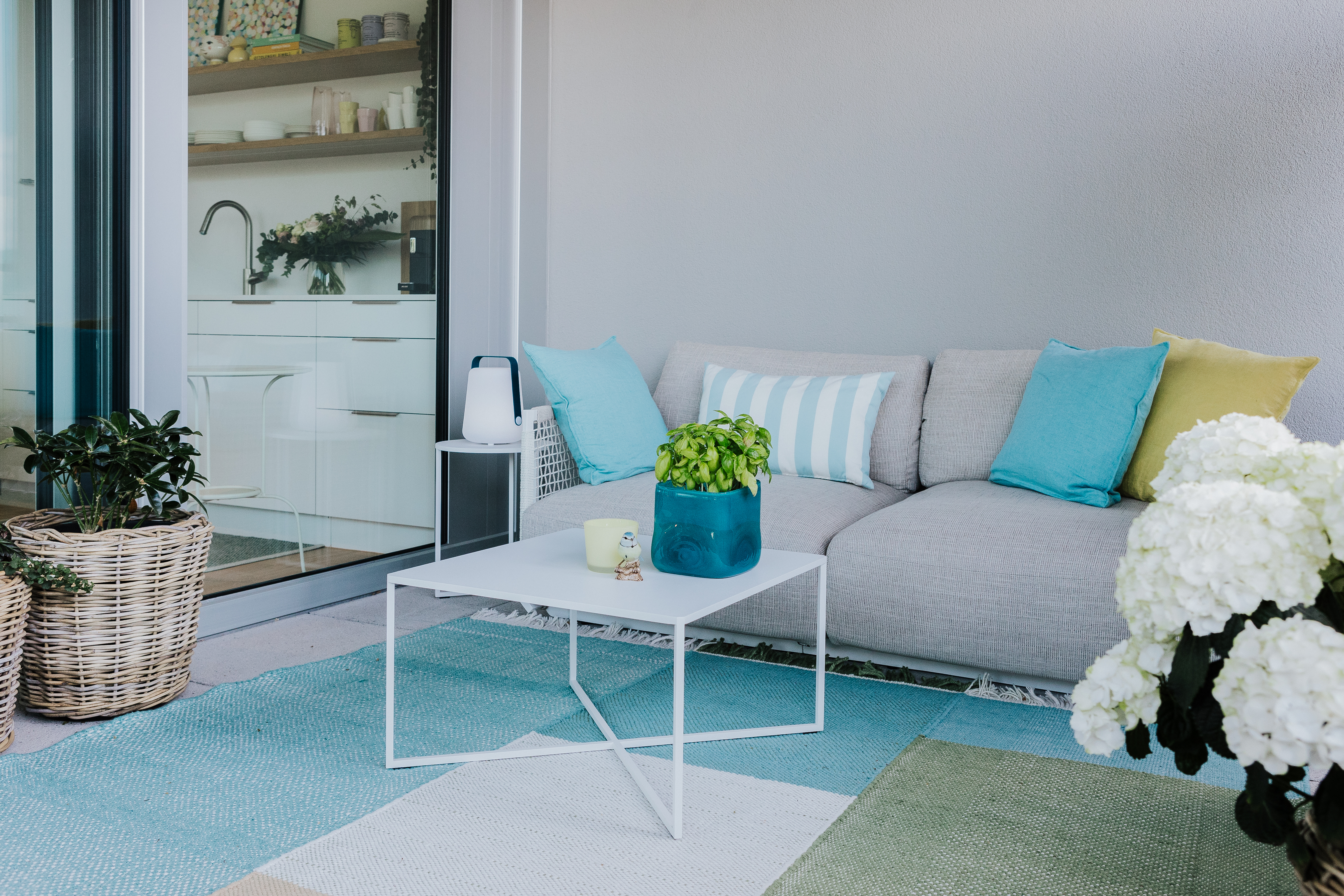 Outdoorsofa mit bunten Kissen, weißem Beistelltisch und Teppichen in Pastelltönen auf Balkon.