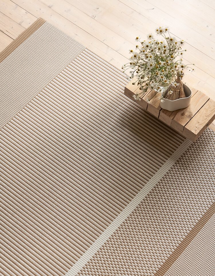 Beiger Teppich mit Streifenstruktur, darauf ein Holztisch mit Vase und Trockenblumen