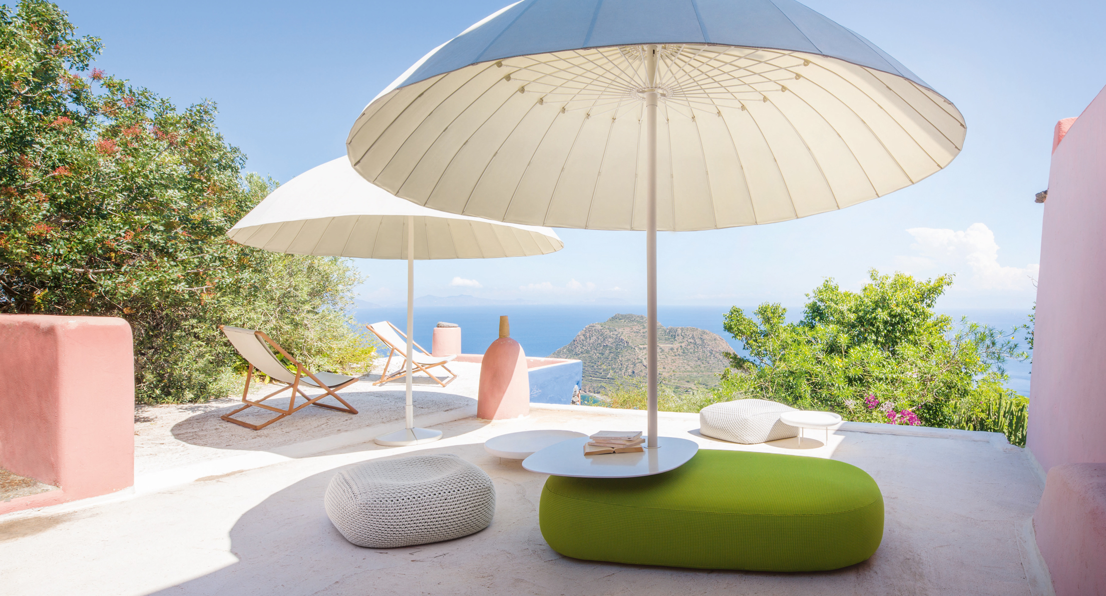 Outdoorbereich mit weißen Schirmen, grüner Sitzinsel, Poufs und Blick aufs Meer im Hintergrund