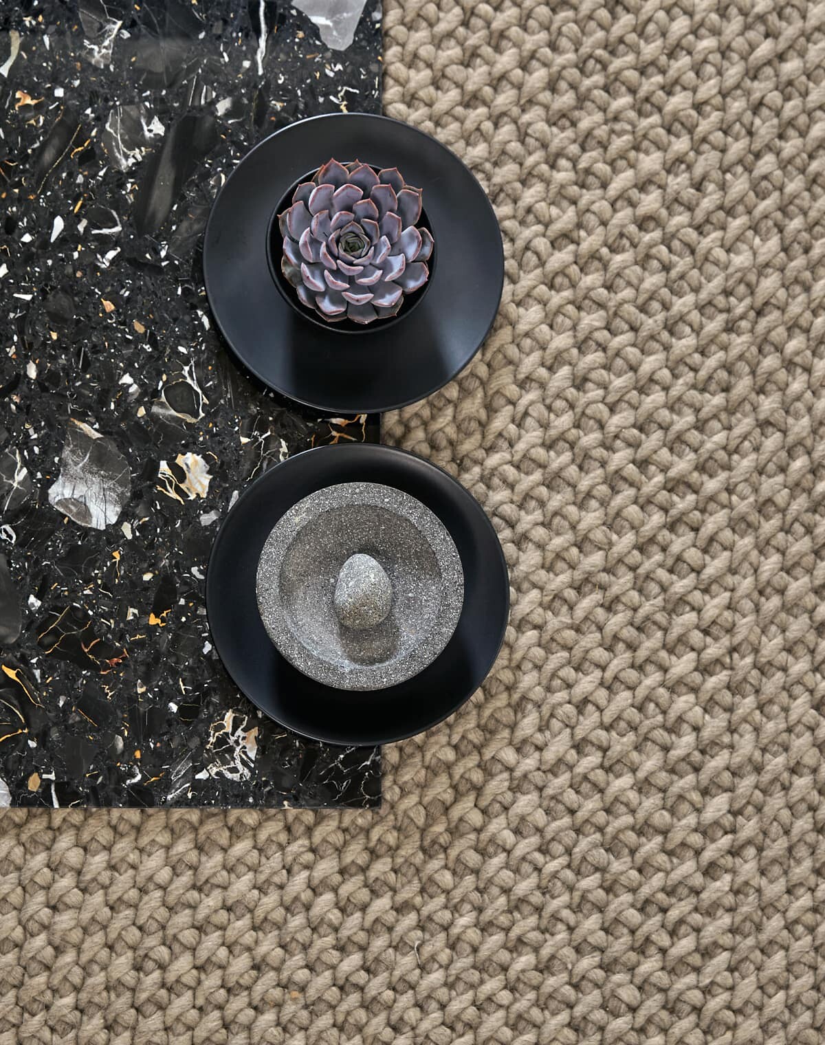 Nahaufnahme eines grob gewebten Teppichs in Taupe mit Deko auf schwarzer Platte