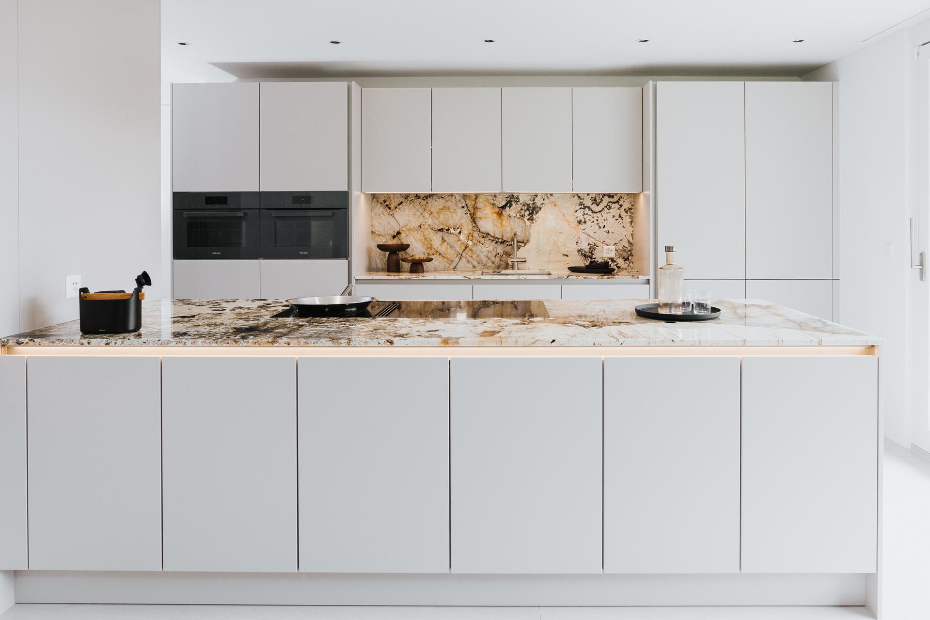 Nolte-Küche mit grifflosen, hellgrauen Fronten und auffälliger Naturstein-Arbeitsfläche mit Wandpaneel.