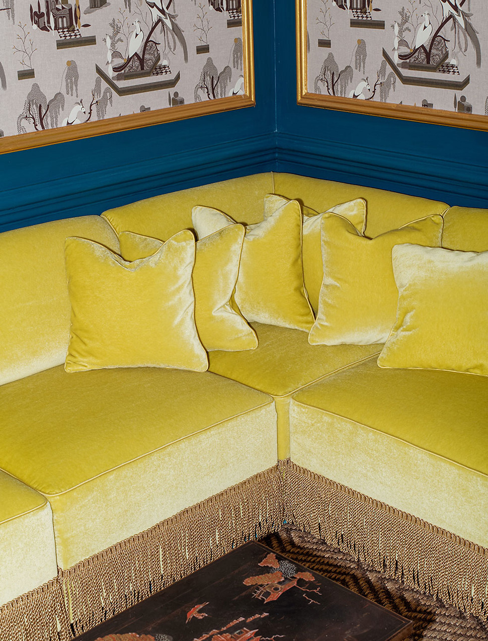 Ecksofa mit gelbem Dedar-Stoff und Fransenborte vor tapezierter Wand mit grafischem Muster