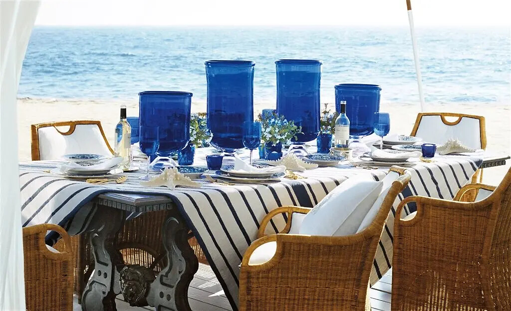 Gedeckter Tisch mit blau-weiß gestreifter Tischdecke und Gläsern am Strand mit Blick aufs Meer