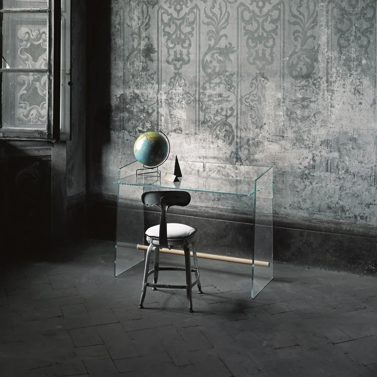 Transparenter Glastisch mit Stuhl in einem dunklen Raum mit ornamentaler Wandstruktur.