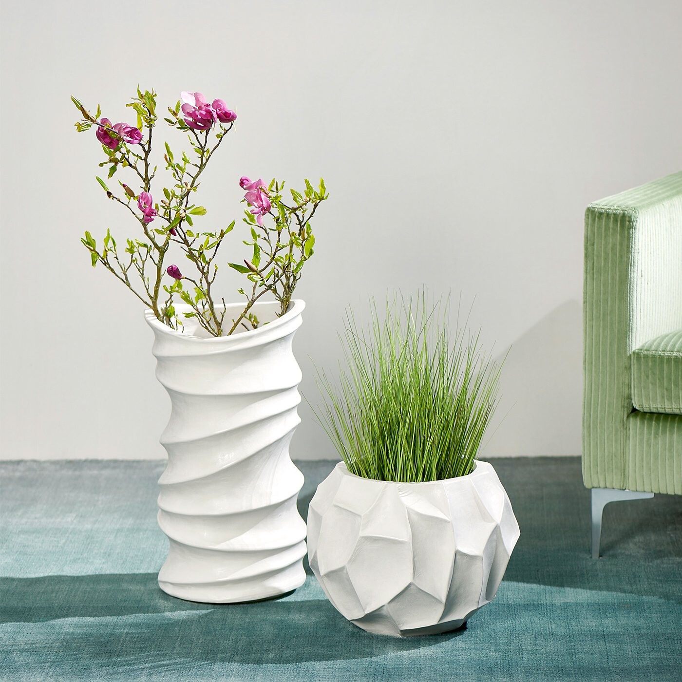 Zwei weiße Pflanzgefäße mit Struktur, eines mit Gräsern, eines mit blühenden Zweigen