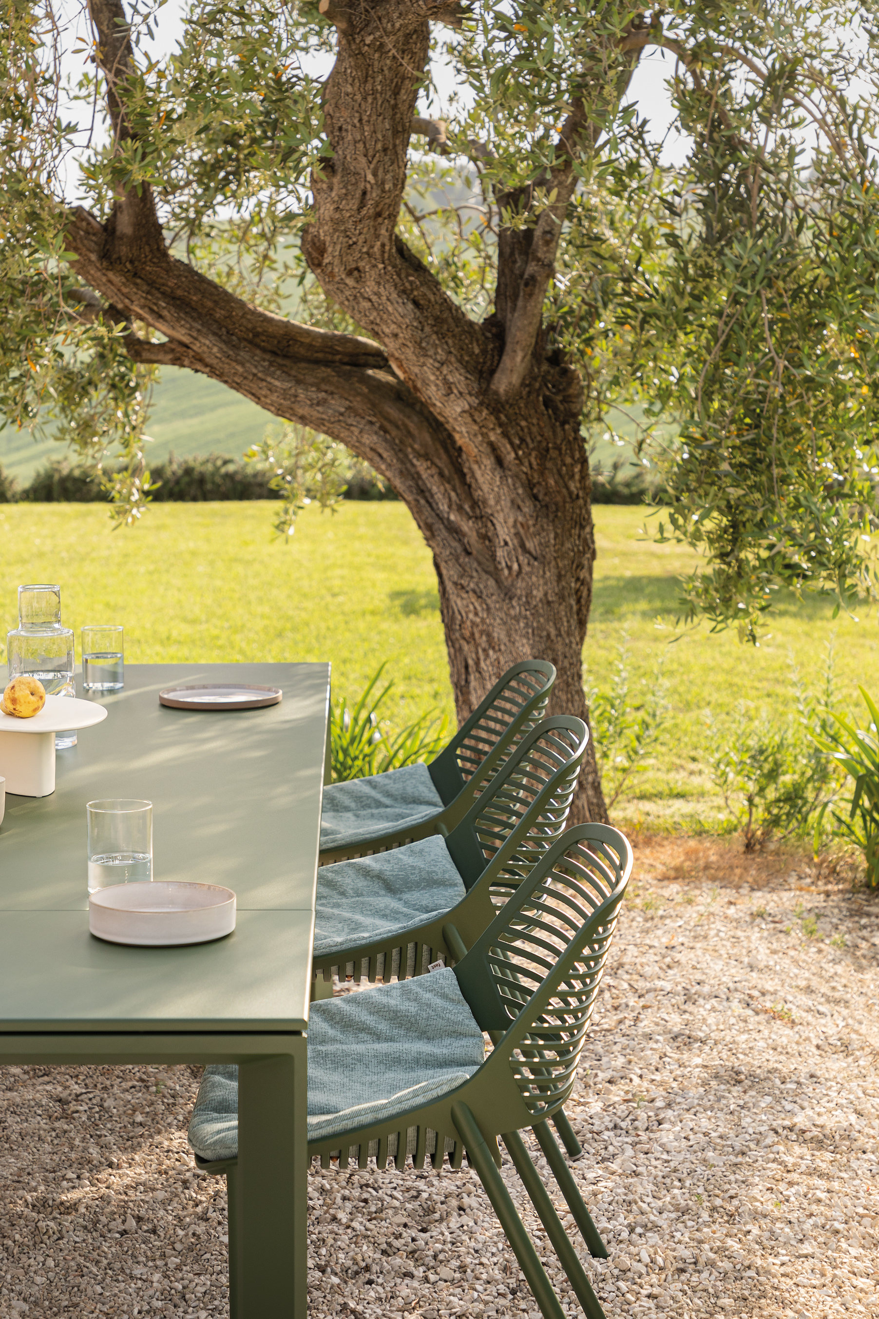 Grüner Outdoor-Esstisch mit stapelbaren Stühlen auf Kiesfläche unter einem alten Olivenbaum