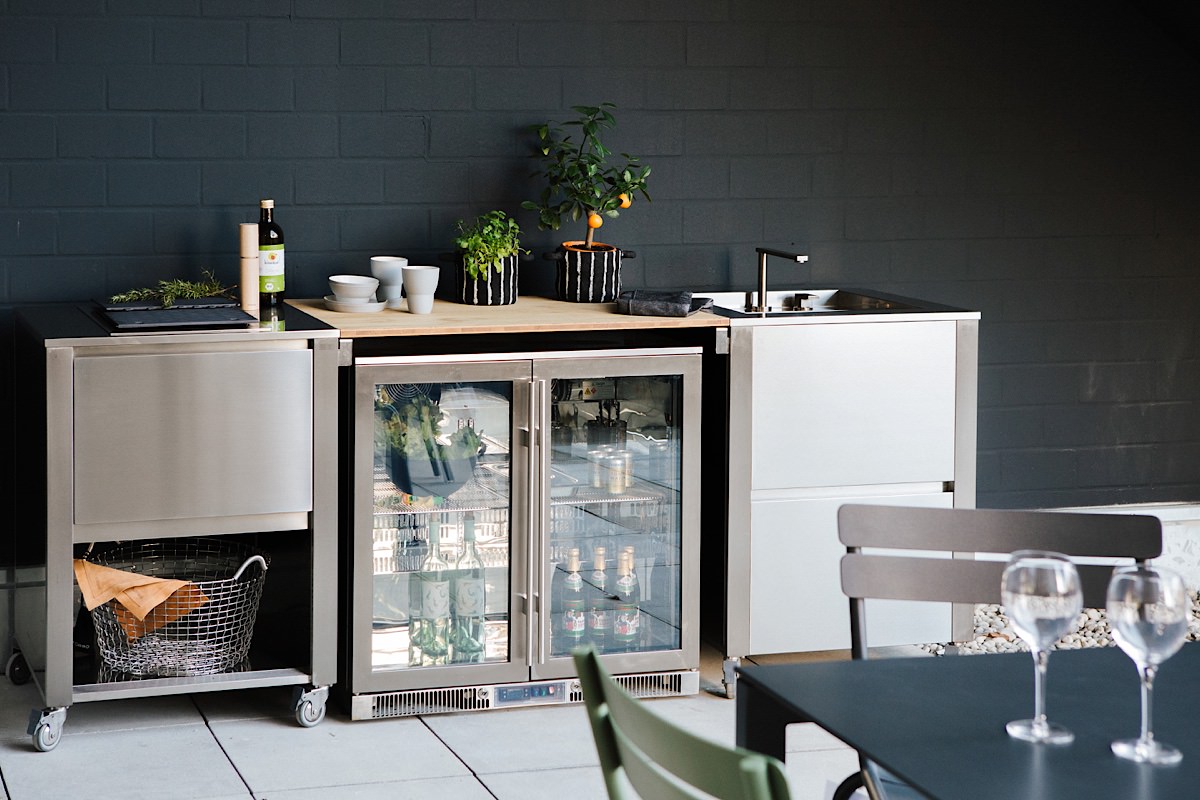 Jokodomus Outdoorküche mit Edelstahlmodulen, integriertem Weinkühlschrank, Spüle und Kräutern auf Arbeitsfläche – platziert auf heller Terrasse.