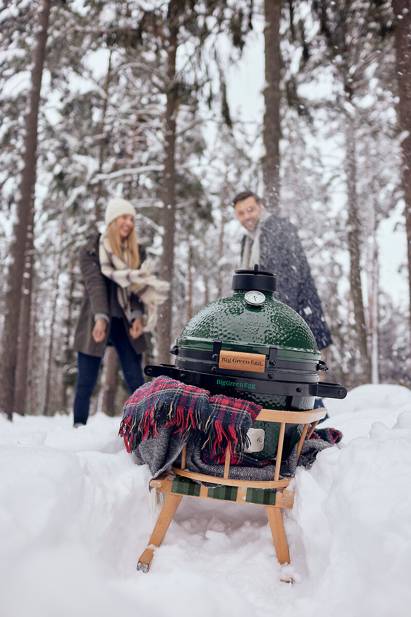 Big Green Egg MiniMax auf Holzschlitten im Schnee, umgeben von Decken und winterlicher Waldkulisse