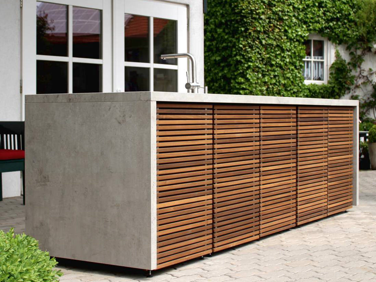Cubic Outdoor Living Küche mit Sichtbetonverkleidung, Holzlamellen und Edelstahlarmatur. Aufgebaut auf einer Terrasse vor begrüntem Haus.