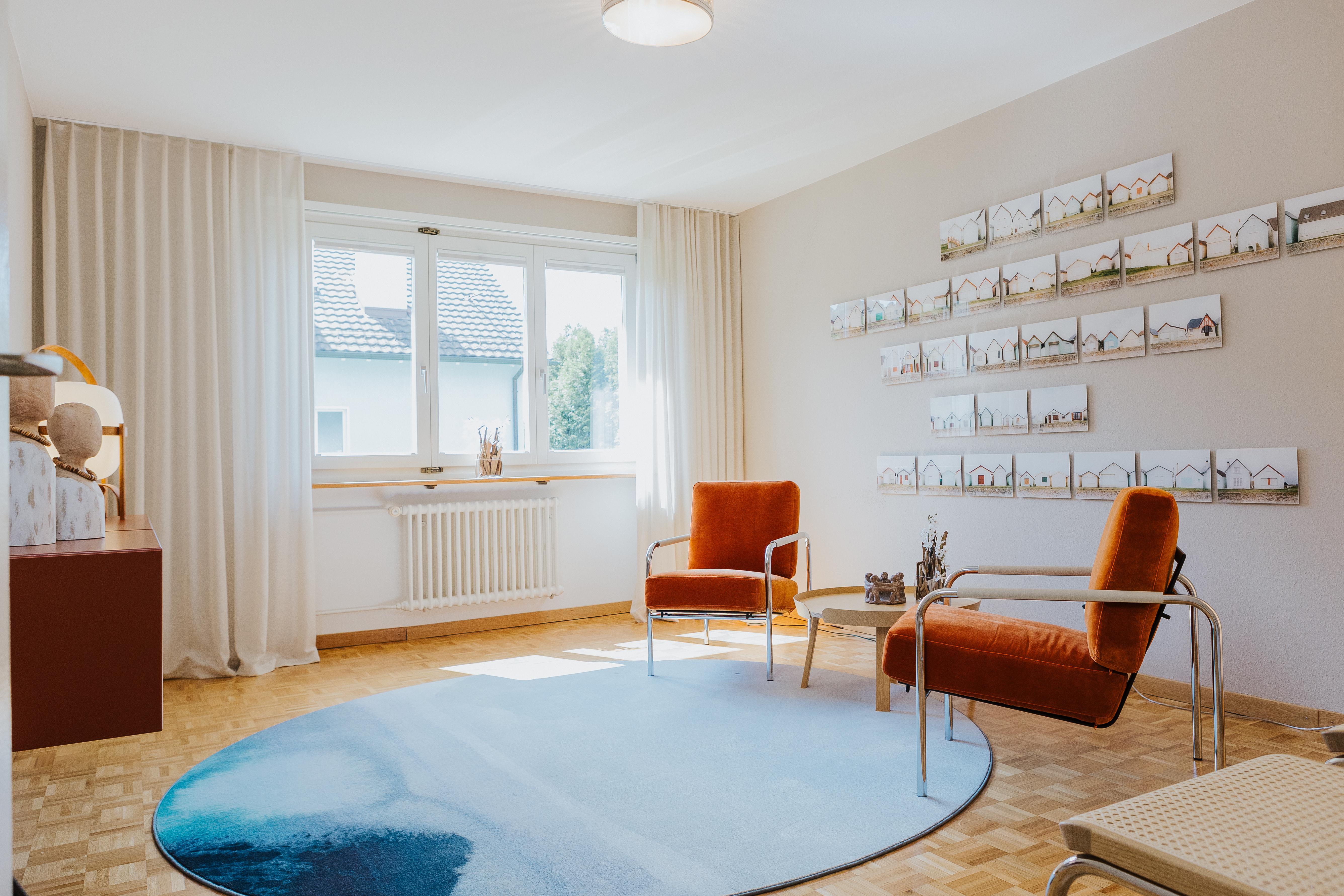 Ruhiger Raum mit zwei orangefarbenen Sesseln, rundem Teppich und Fotoreihe an der Wand.