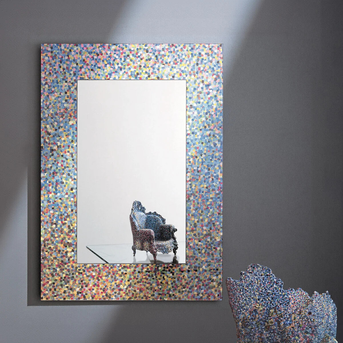 Spiegel mit farbigem, mozaikartigem Rahmen vor grauer Wand, Stuhl im Spiegelbild.