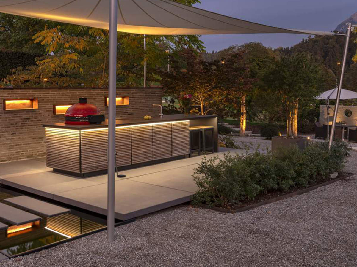 Beleuchtete Outdoorküche von Cubic Outdoor Living in der Dämmerung. Holzlamellen, roter Grill, Sonnensegel und stilvolle Gartenarchitektur.