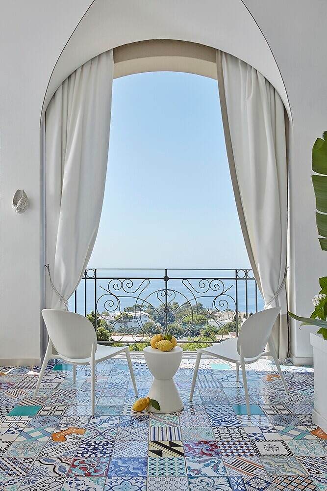 Zwei weiße Stühle vor Balkon mit gemustertem Boden und Blick aufs Meer