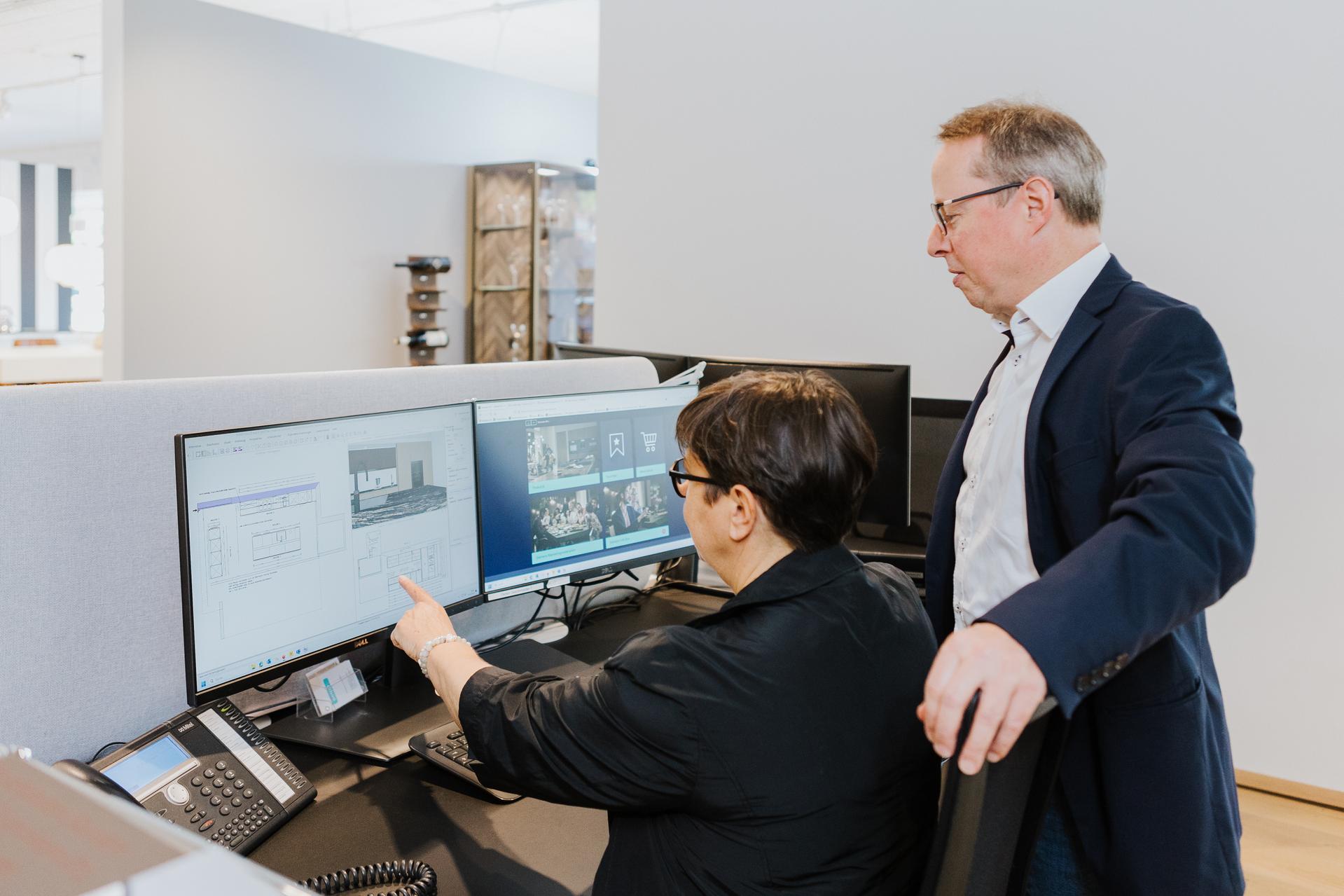 Zwei Küchenberater im Gespräch vor Monitoren mit 3D-Planung und Küchenansichten