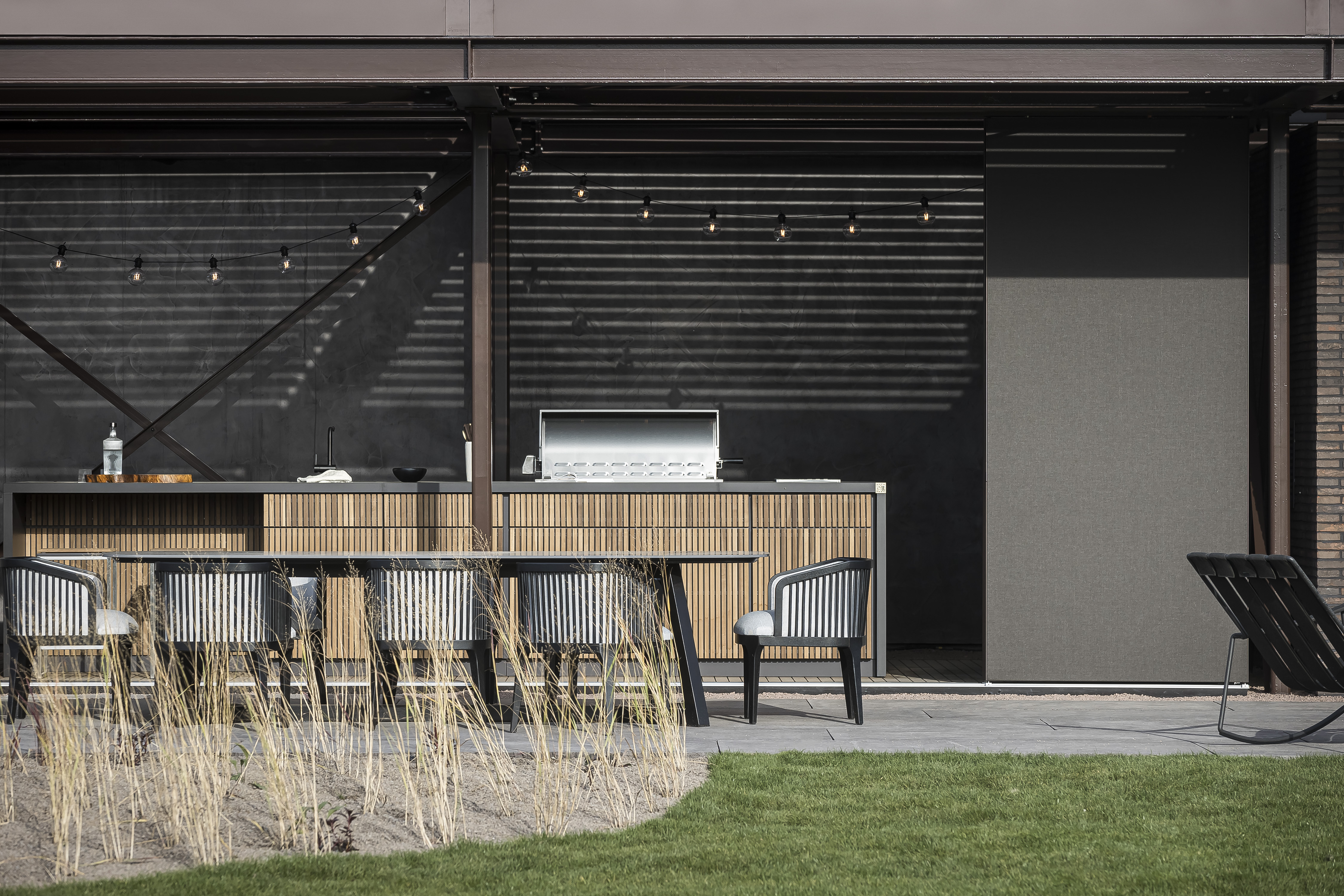 Outdoor-Küche von Cubic Outdoor Living unter einer überdachten Terrasse mit dunkler Rückwand und Barbereich. Davor Esstisch mit Flechtstühlen und Blick auf eine gepflegte Rasenfläche.