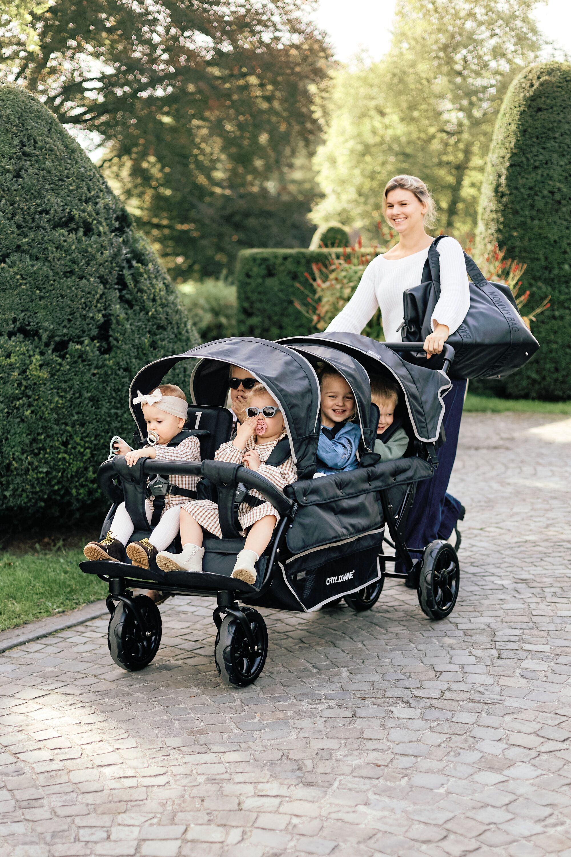 Frau schiebt Kinderwagen mit sechs kleinen Kindern auf Weg durch eine gepflegte Parkanlage
