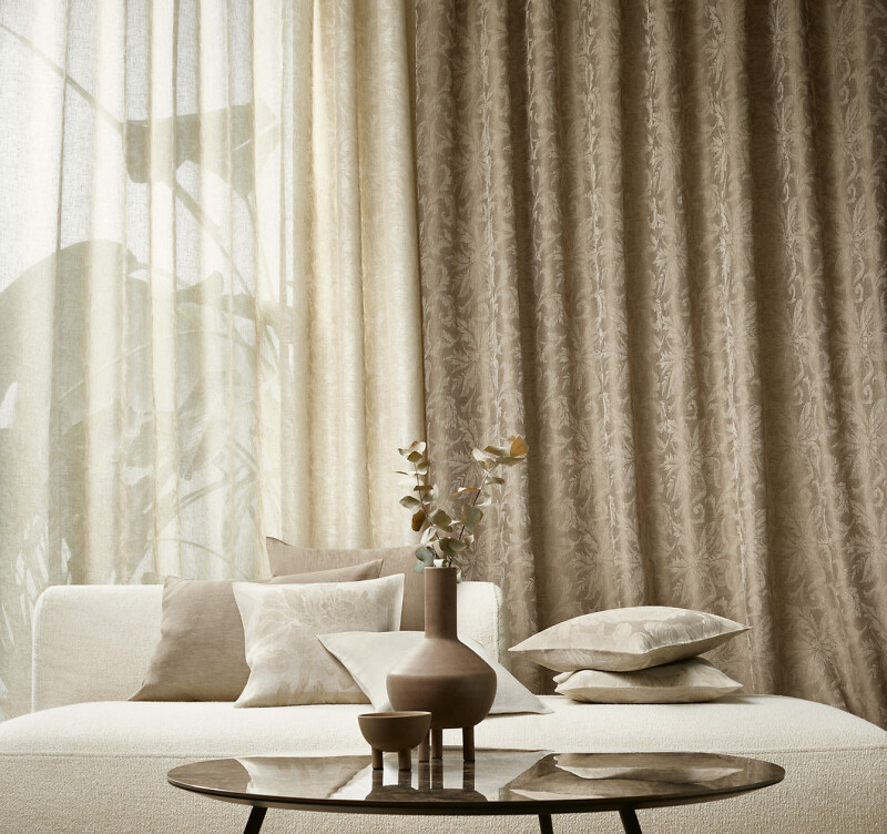 Zwei Lagen beigefarbener Vorhänge hinter hellen Zierkissen auf einem Sofa