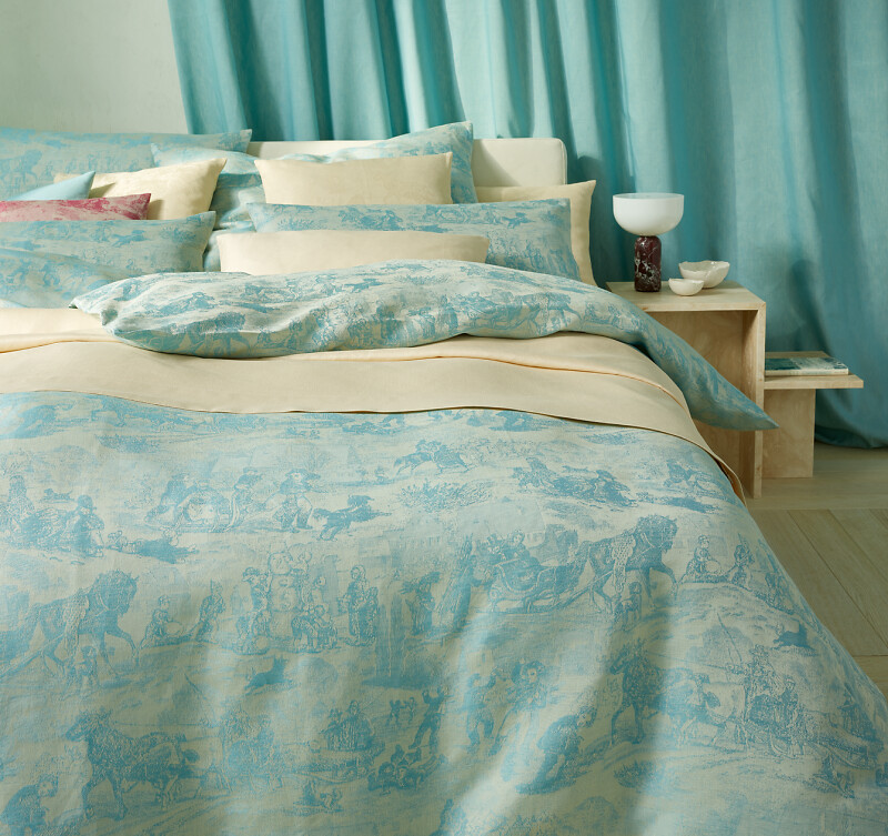 Bett mit mehrlagiger Bettwäsche in Blau und Beige vor türkisfarbenem Vorhang