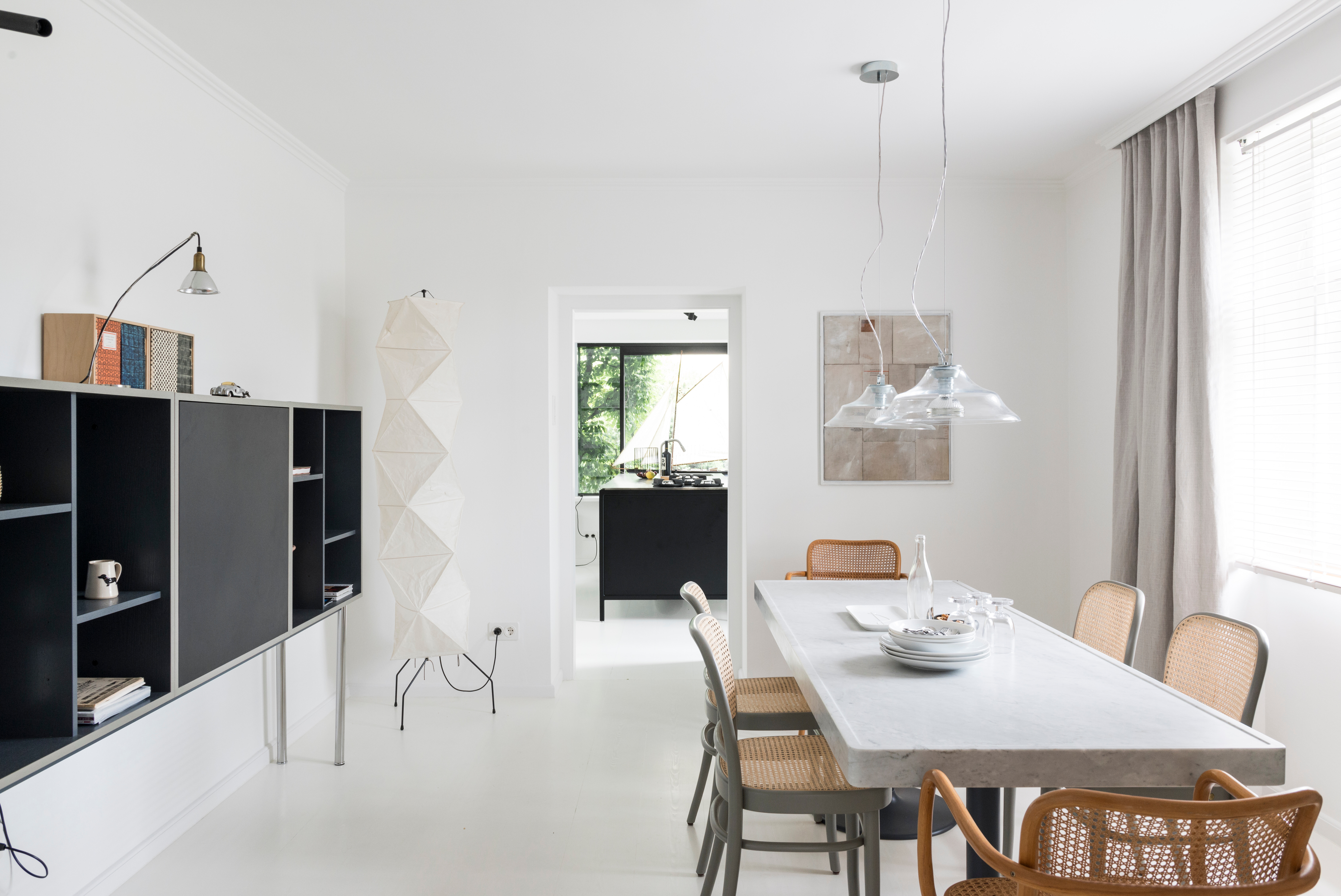 Heller Essbereich mit schlichten Holzmöbeln, Pendelleuchten und Blick in eine offene schwarze Küche.