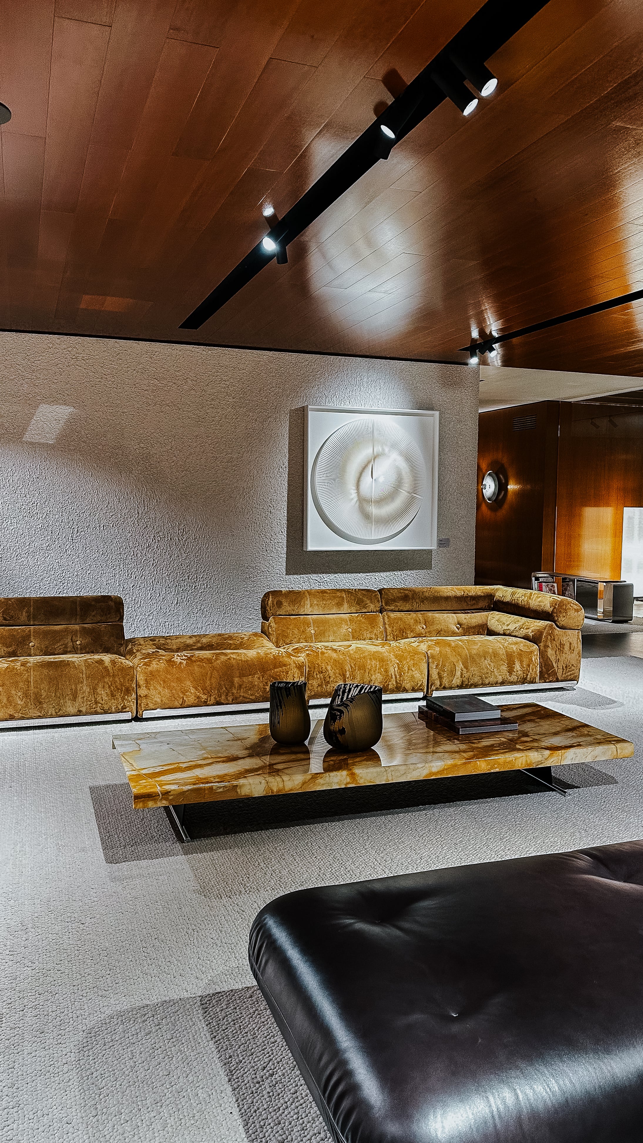 Sofalandschaft in cognacfarbenem Leder auf der Il Salone del Mobile 2025. Die Szene umfasst einen Natursteintisch, eine plastisch strukturierte Wand mit Kunstobjekt und eine Decke mit dunkler Holzverkleidung.