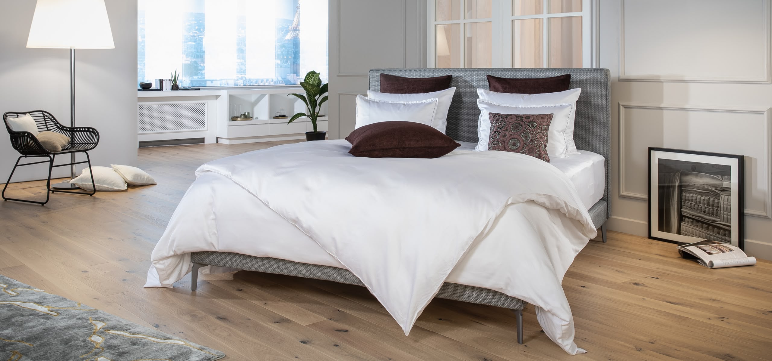 Boxspringbett mit grauem Kopfteil, weißer Bettwäsche und dunklen Kissen auf Holzfußboden