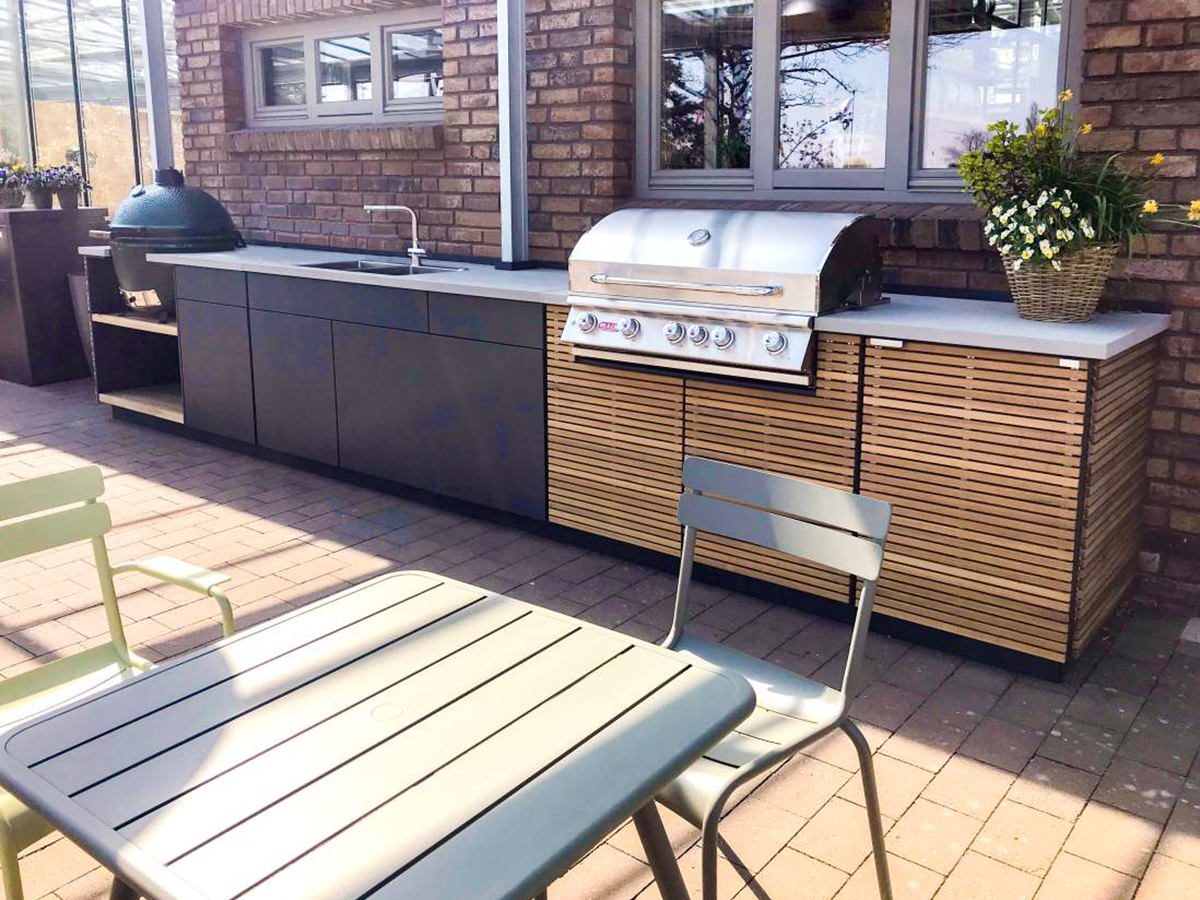 Outdoorküche von Cubic Outdoor Living mit Edelstahlgrill, Keramikspüle und Holzlamellenfront vor Backsteinwand. Im Vordergrund ein Tisch.